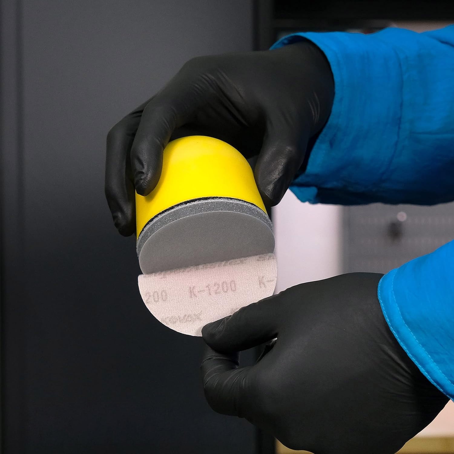 Person wearing black gloves holding a yellow and gray sanding disc against a dark background