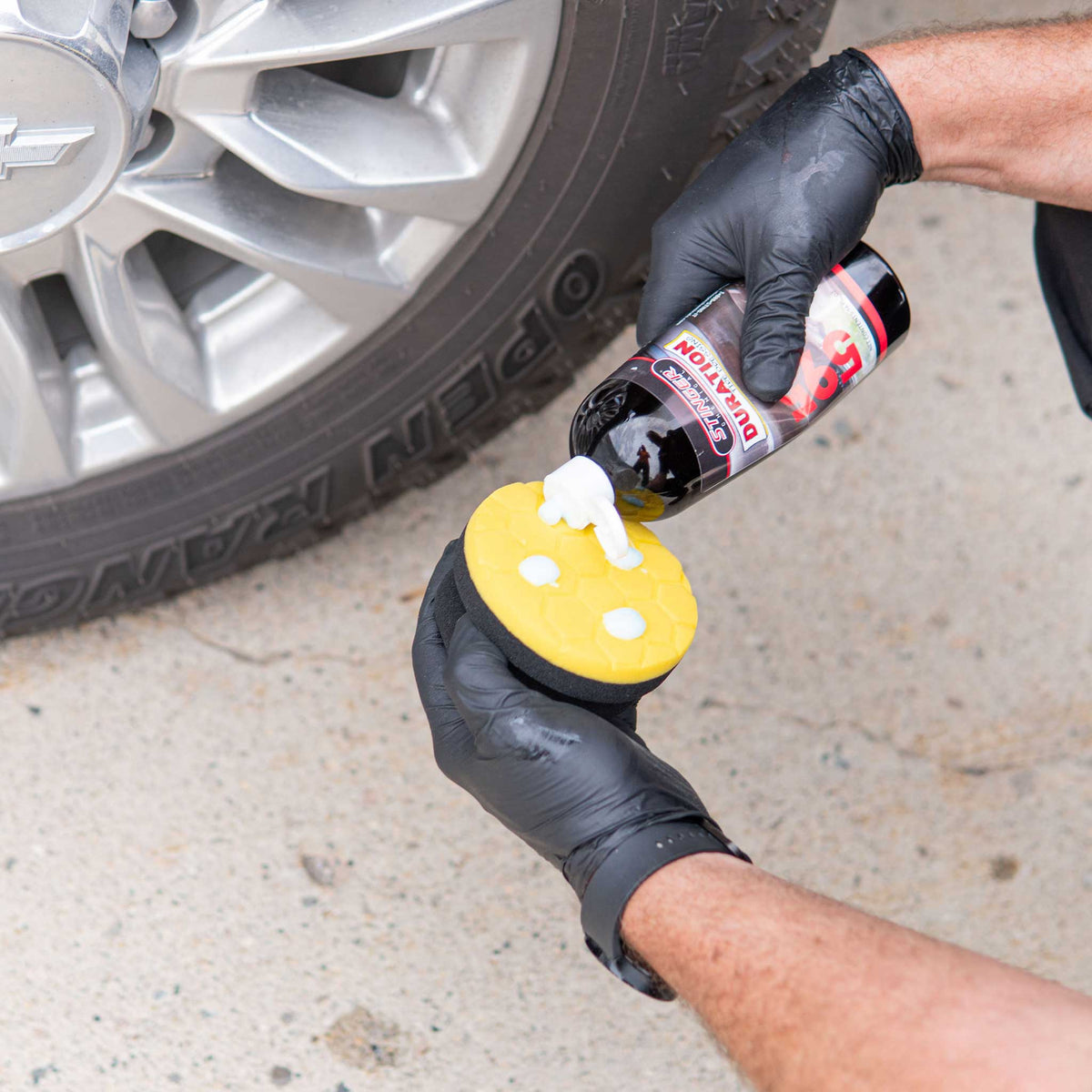 A person wearing black gloves Applying Duration Tire Dressing from a bottle onto a yellow hex tire dressing applicator sponge near a car tire on a concrete surface