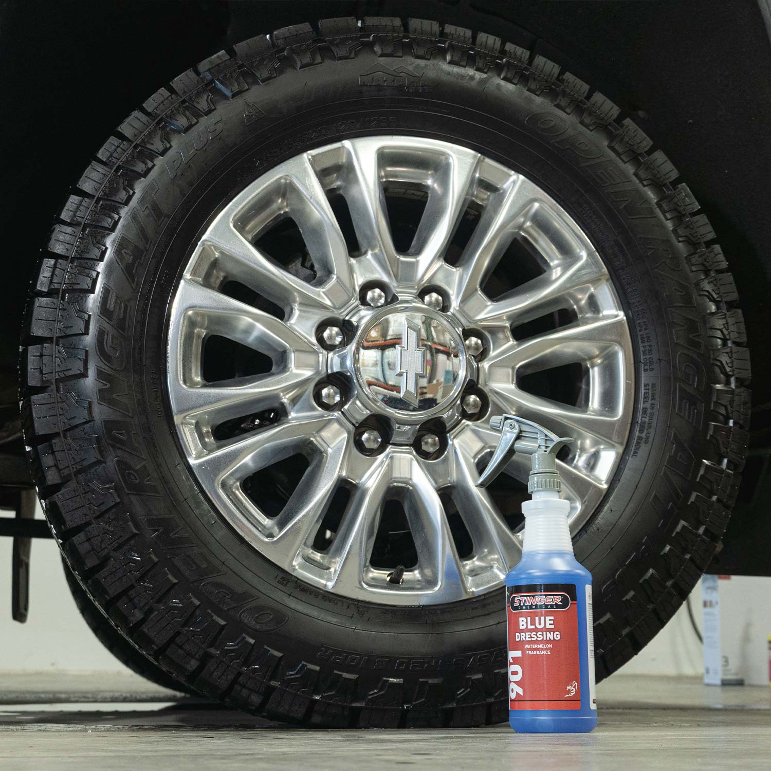 Car tire with chrome rim and a bottle of tire dressing on a shop floor.