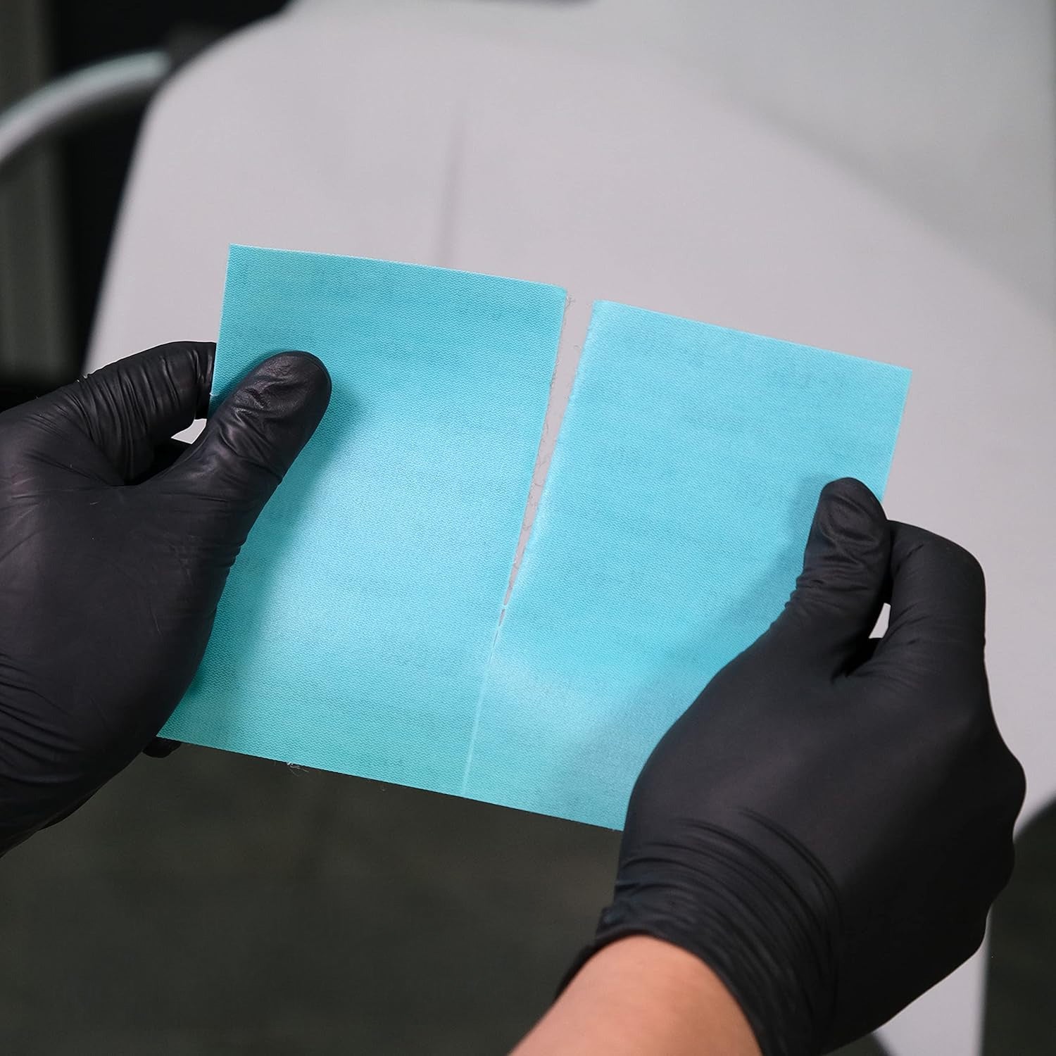 Hands wearing black gloves tearing apart two blue sandpaper patches against a neutral background
