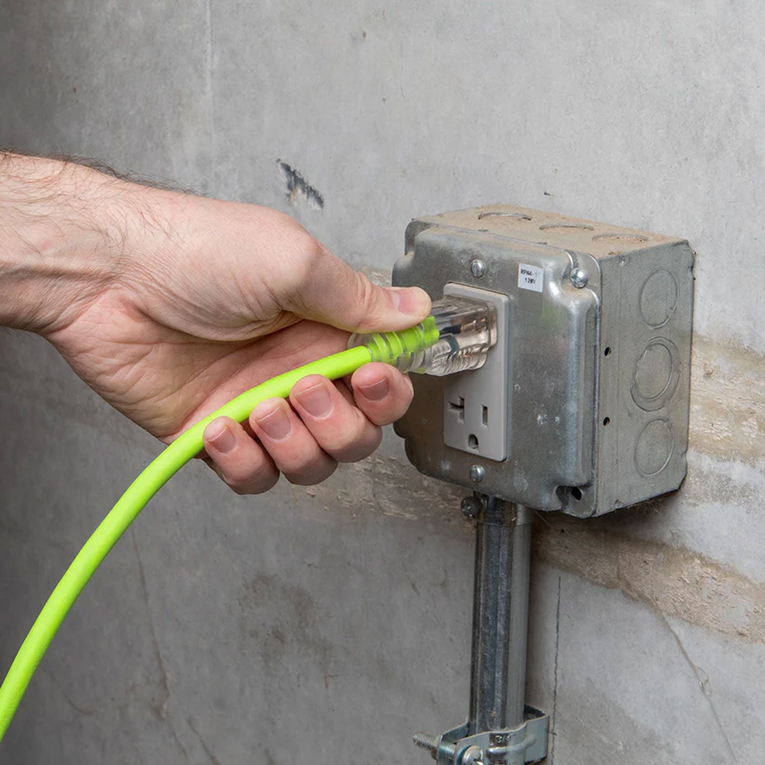 Hand plugging a green cable into an electrical outlet on a concrete wall.