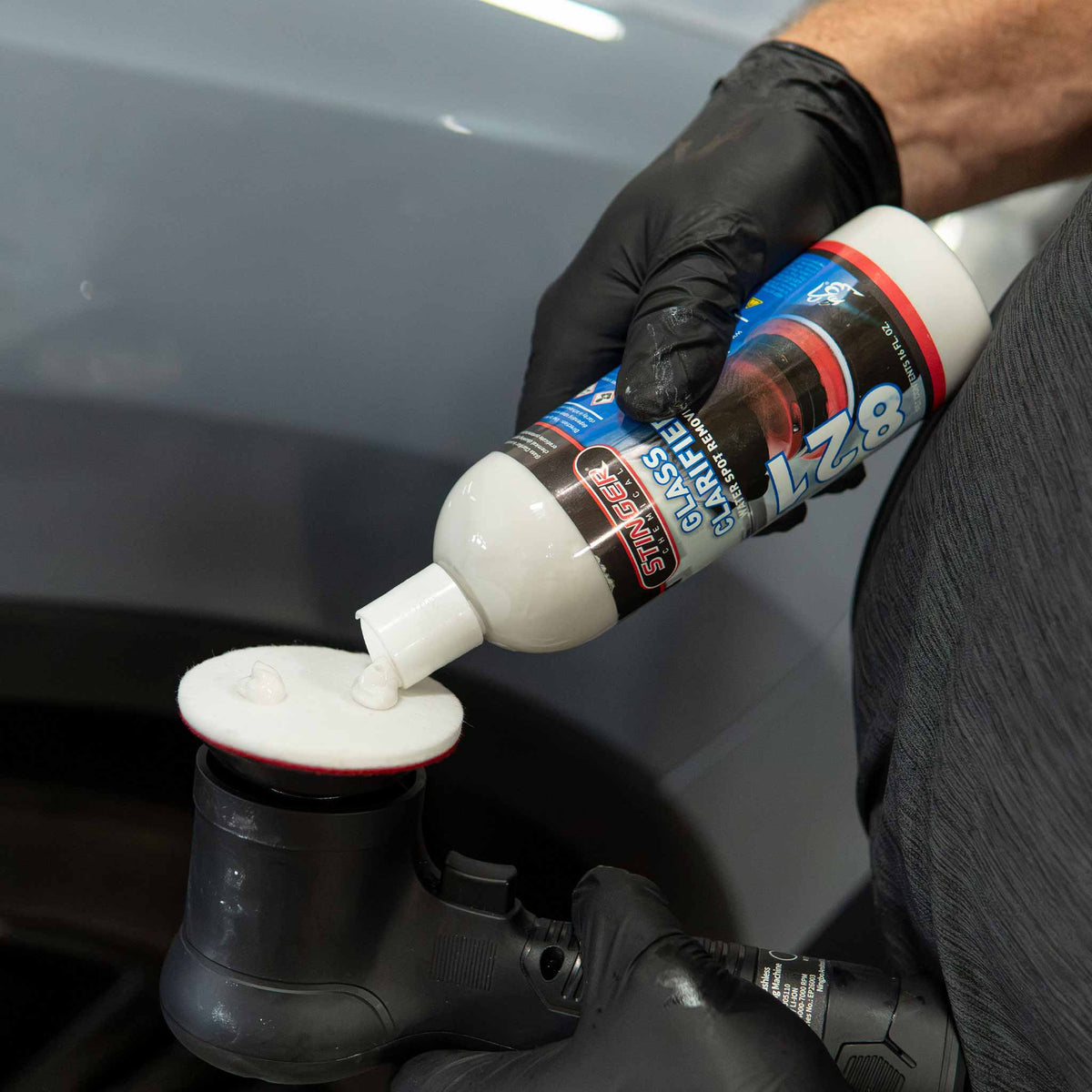 A person wearing a black glove squeezes white polish from a bottle labeled Glass Clarifier onto a white rayon glass pad, with a gray car body in the background.