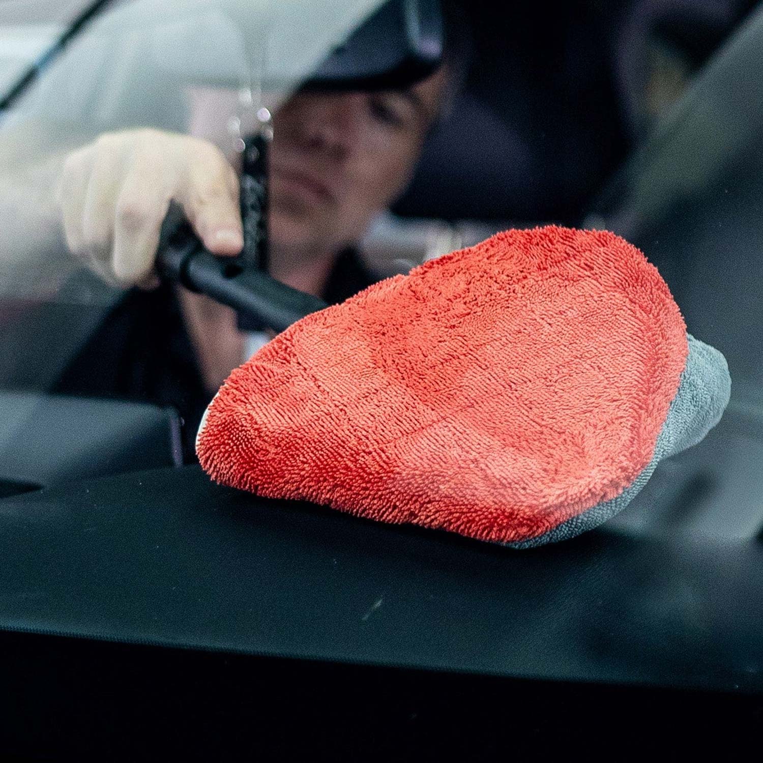 Red microfiber towel on a car surface with a person cleaning the car in the background.