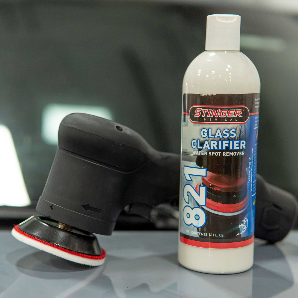 A bottle of Stinger Chemicals 821 Glass Clarifier water spot remover stands next to a black Barjack Raven orbital polisher with a red and white foam pad on a glossy surface, with a blurred car interior in the background.