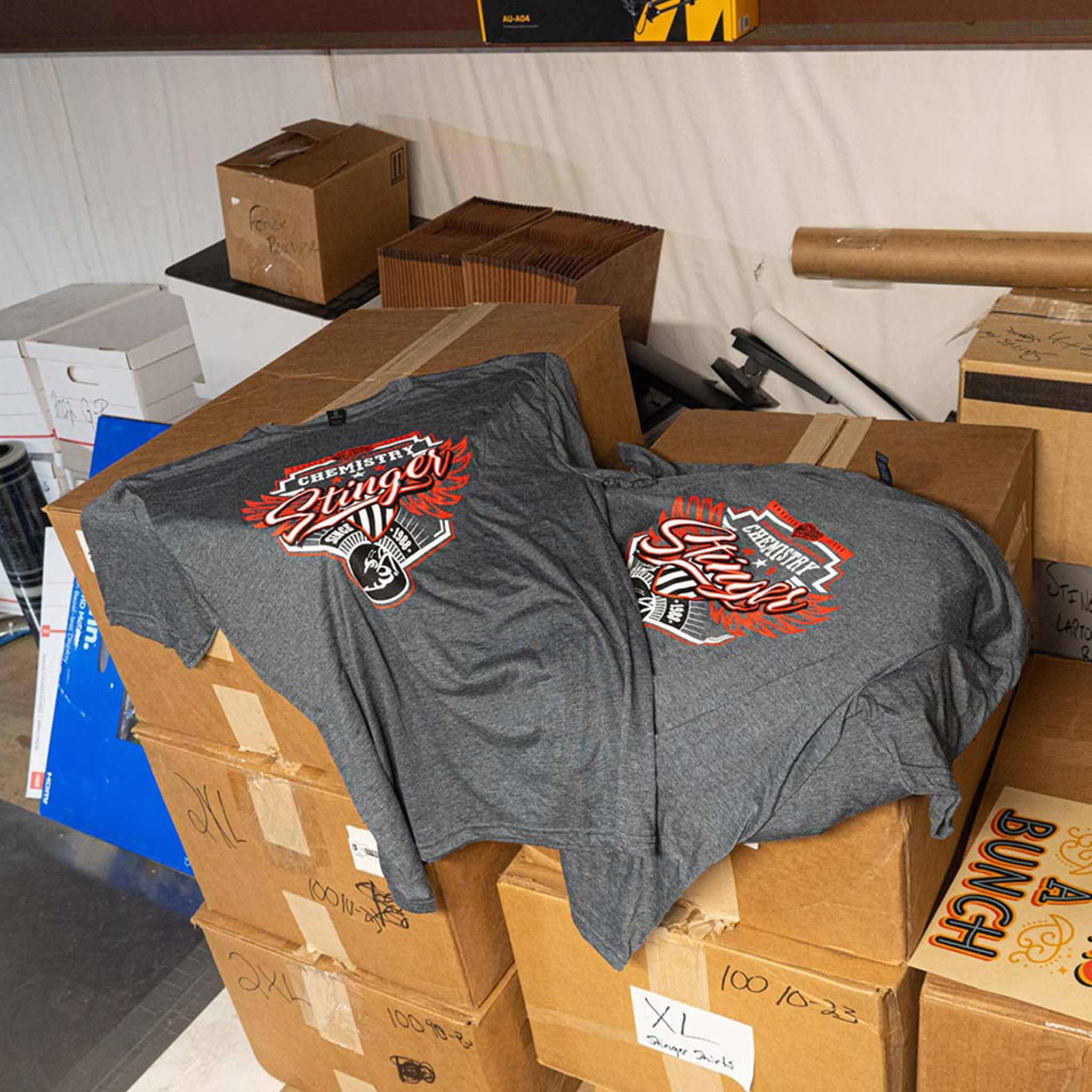 Gray t-shirt with a logo on a stack of cardboard boxes in a storage room.