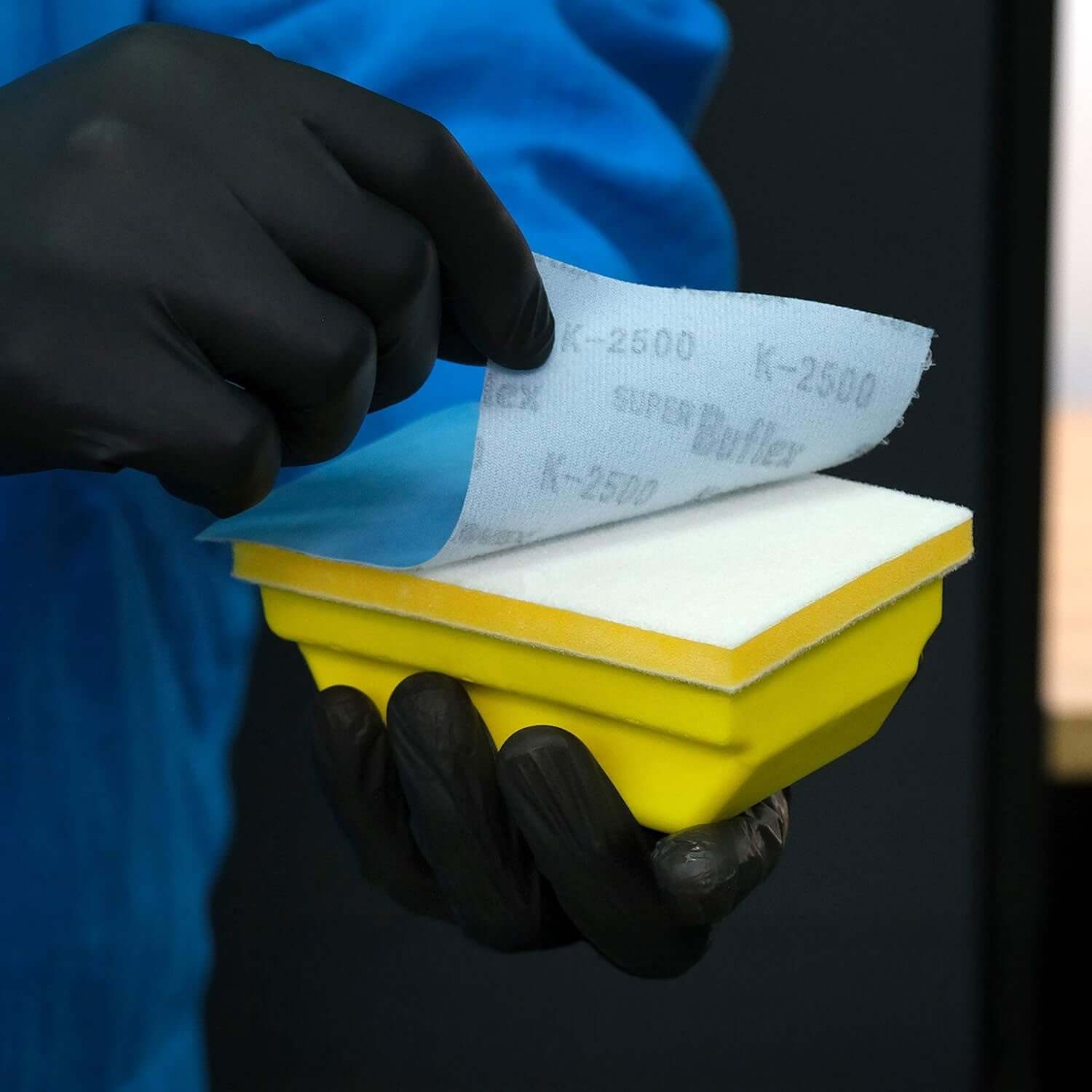 Person wearing black gloves holding a yellow sanding block with Buflex K-2500 sandpaper.
