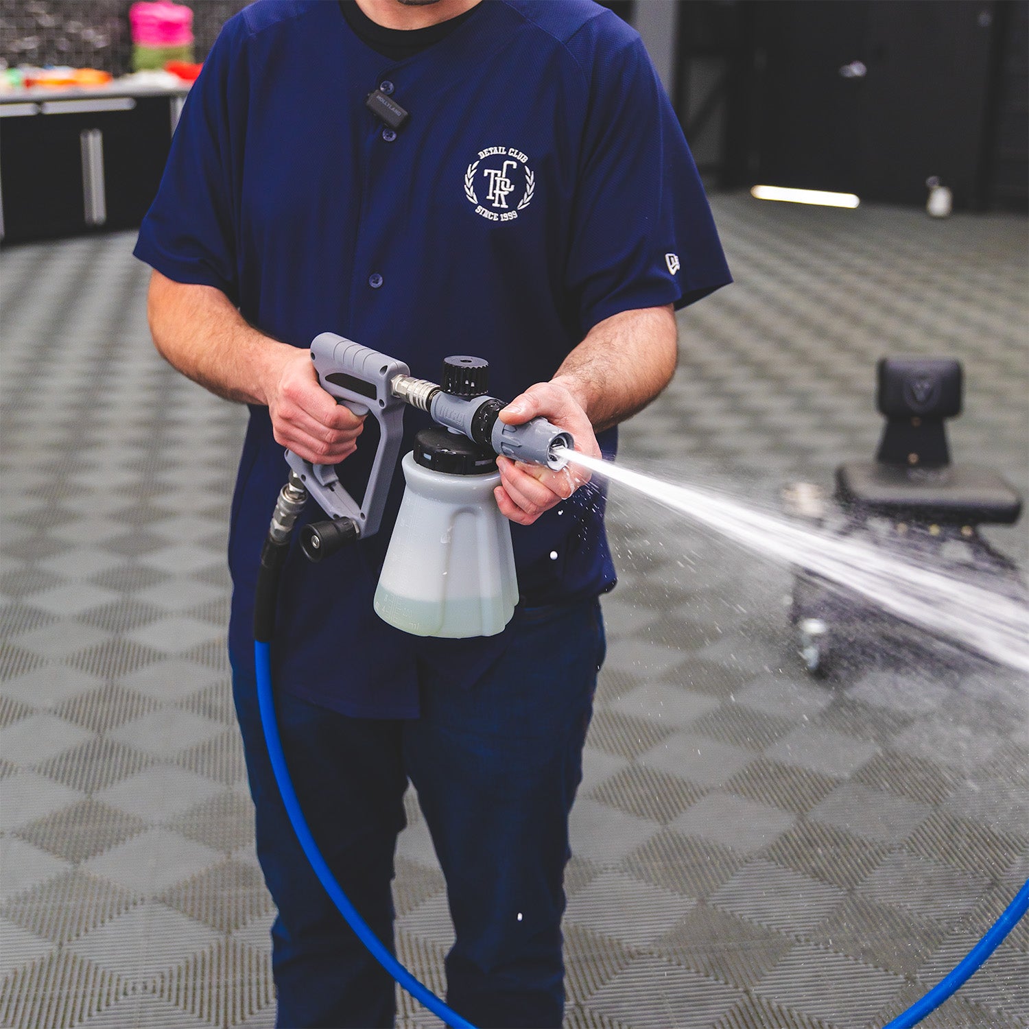 A person in a navy blue scrub shirt holds a foaming spray gun connected to a hose, spraying a fine mist in a clean indoor setting with a gray checkered floor.