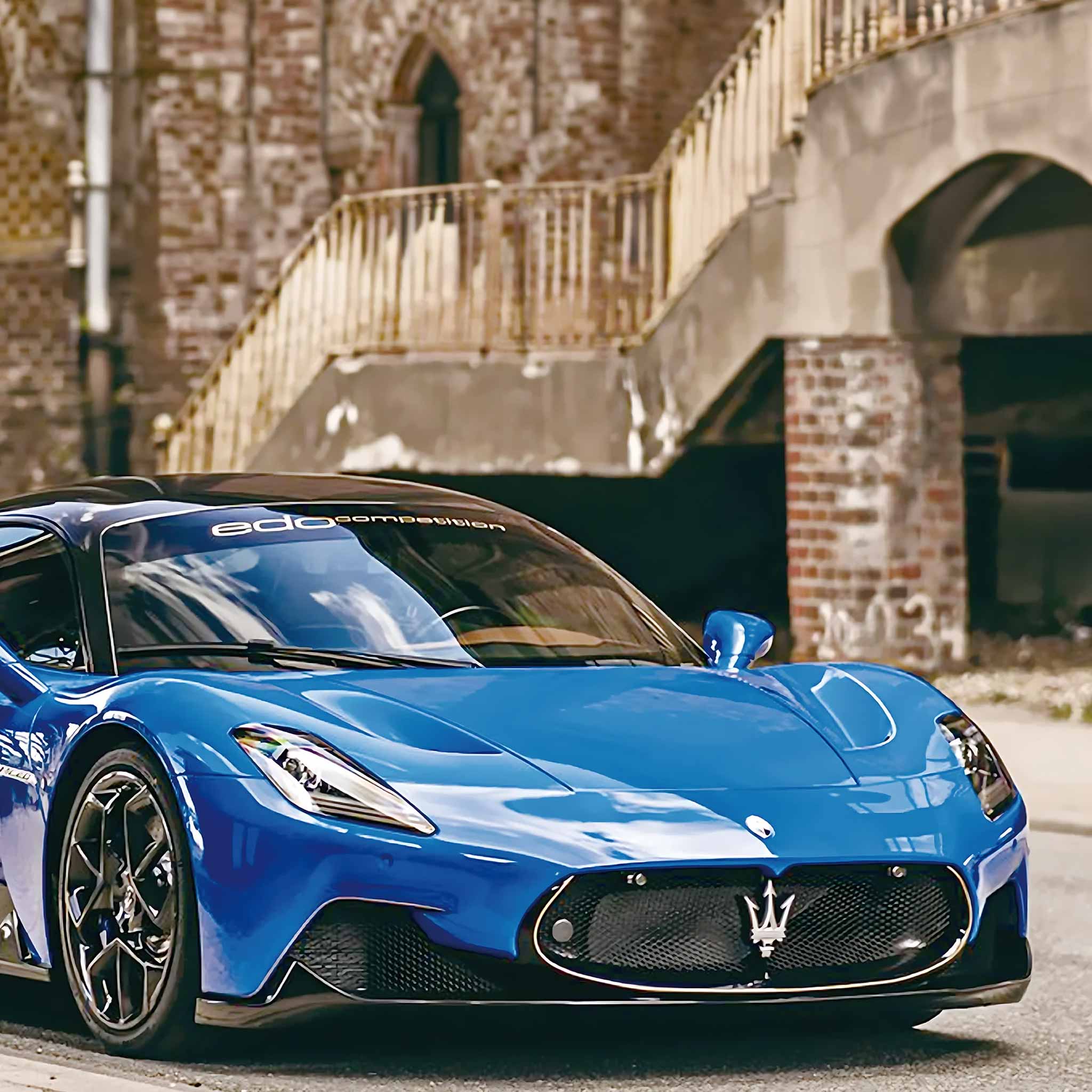 A bright blue Maserati hypercar with black wheels and a black windshield, photographed in motion on a street with an urban brick bridge background.