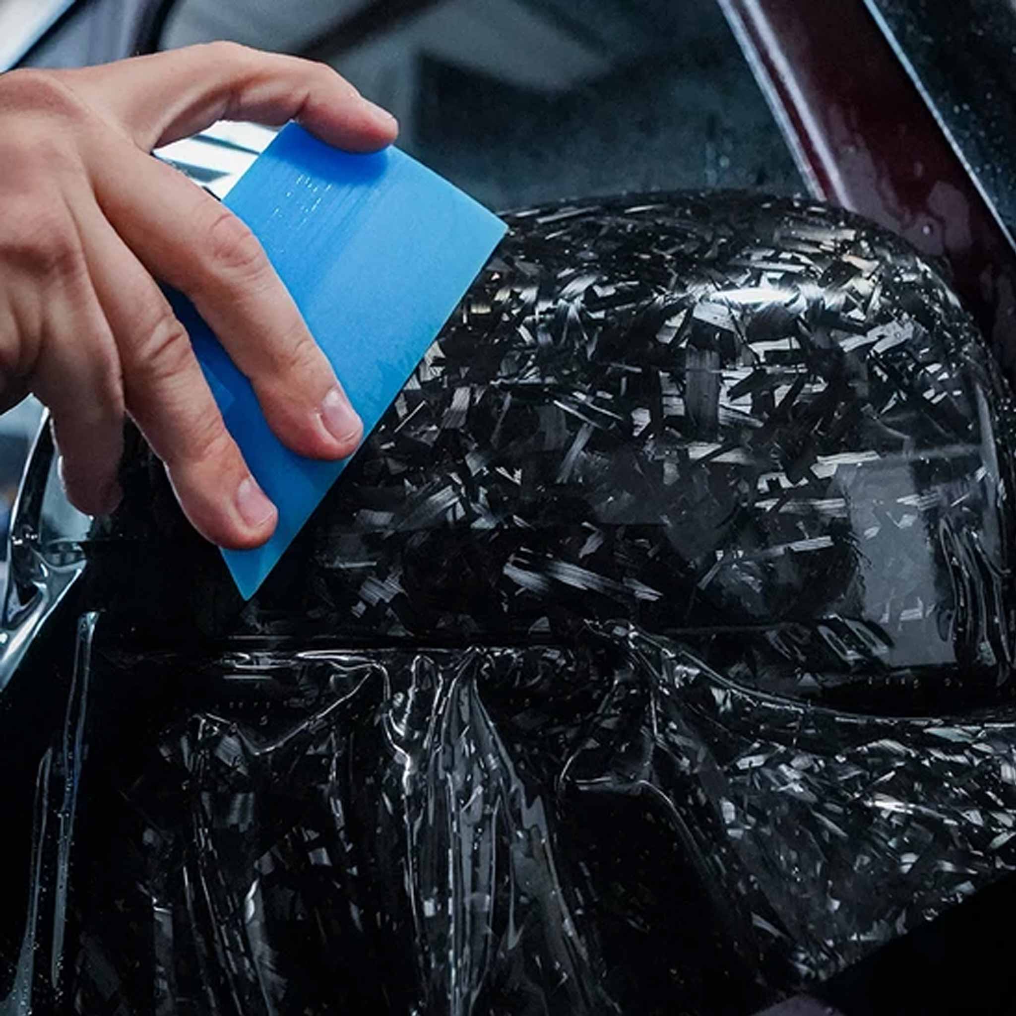 A close-up of a hand using a blue squeegee to apply a clear protective film over a black car part.