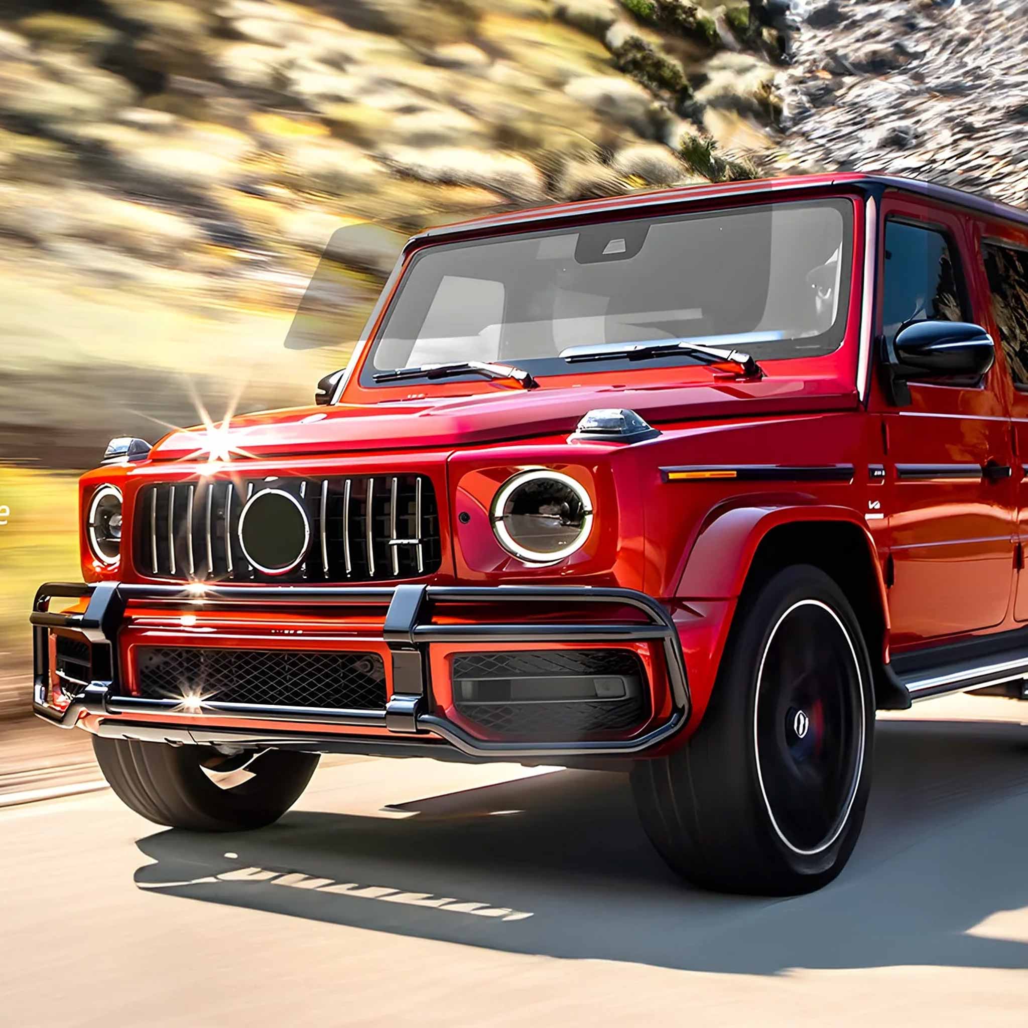 A red Mercedes-Benz G-Class SUV is shown driving on a road with a blurred rocky mountain background. The front grille features the Mercedes-Benz logo, and the vehicle has black wheels and a black front bumper guard.