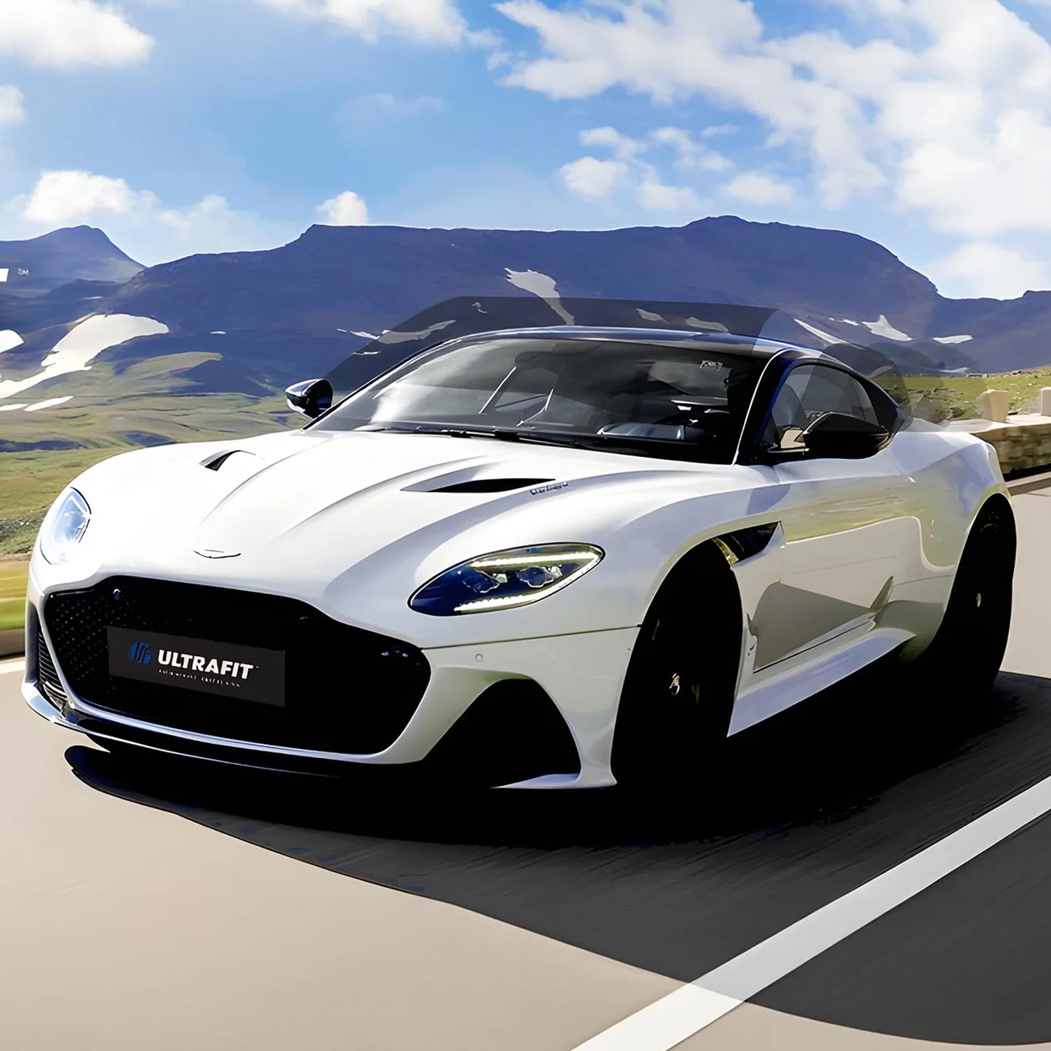 A white Aston Martin DBS with an ULTRAFIT black window tint on its glass, roof, and side panels, shown driving on a road with mountains in the background.