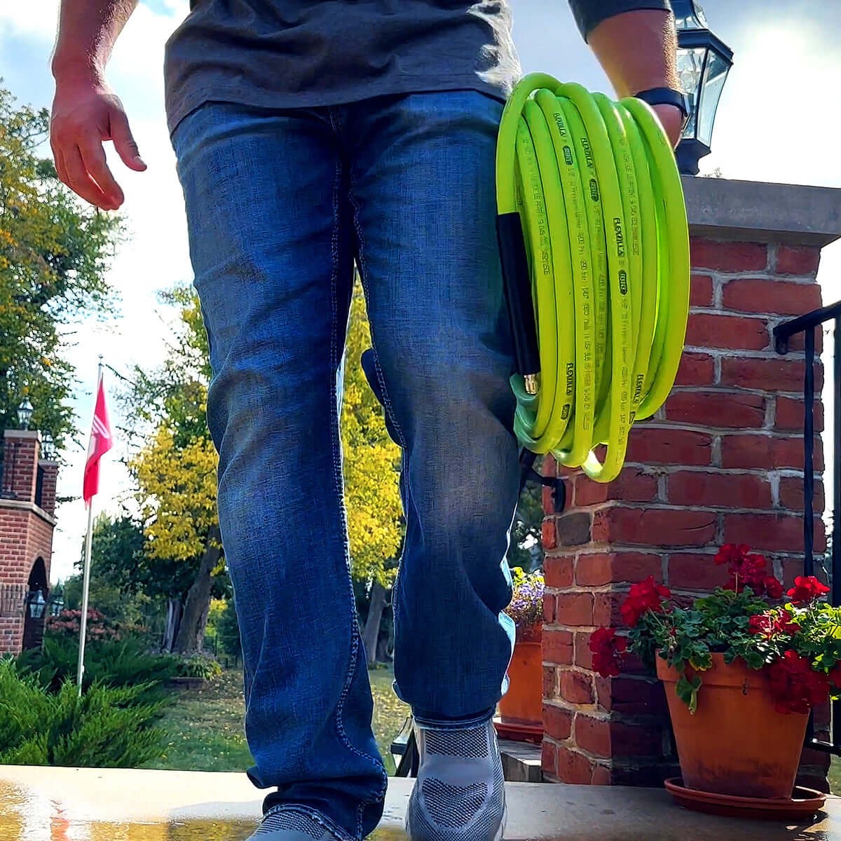 Person holding a coiled green pressure washer hose with a brick wall and potted plants in the background