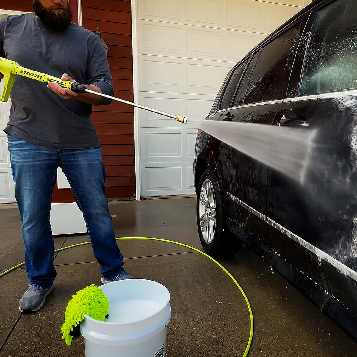 Person washing a black car with a pressure washer in front of a garage.
