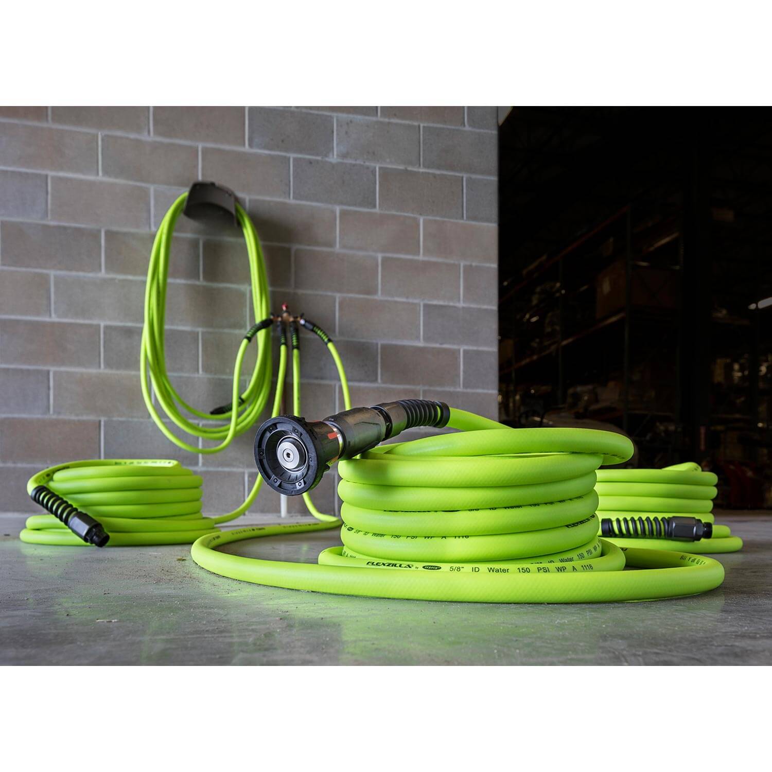 Green garden hose with nozzle on a concrete floor against a brick wall.