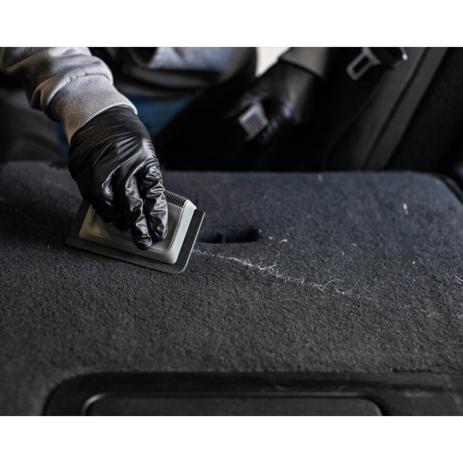 Person cleaning pet hair off of a car's black carpet with a brush and black gloves.