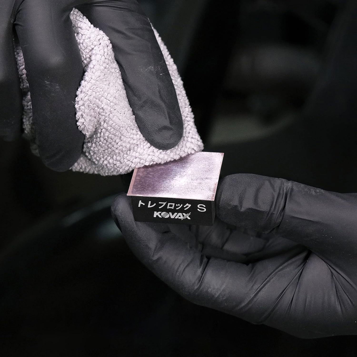 Black Glove holding a KOVAX toleblock product removing slurry with a microfiber towel on a dark blurred background