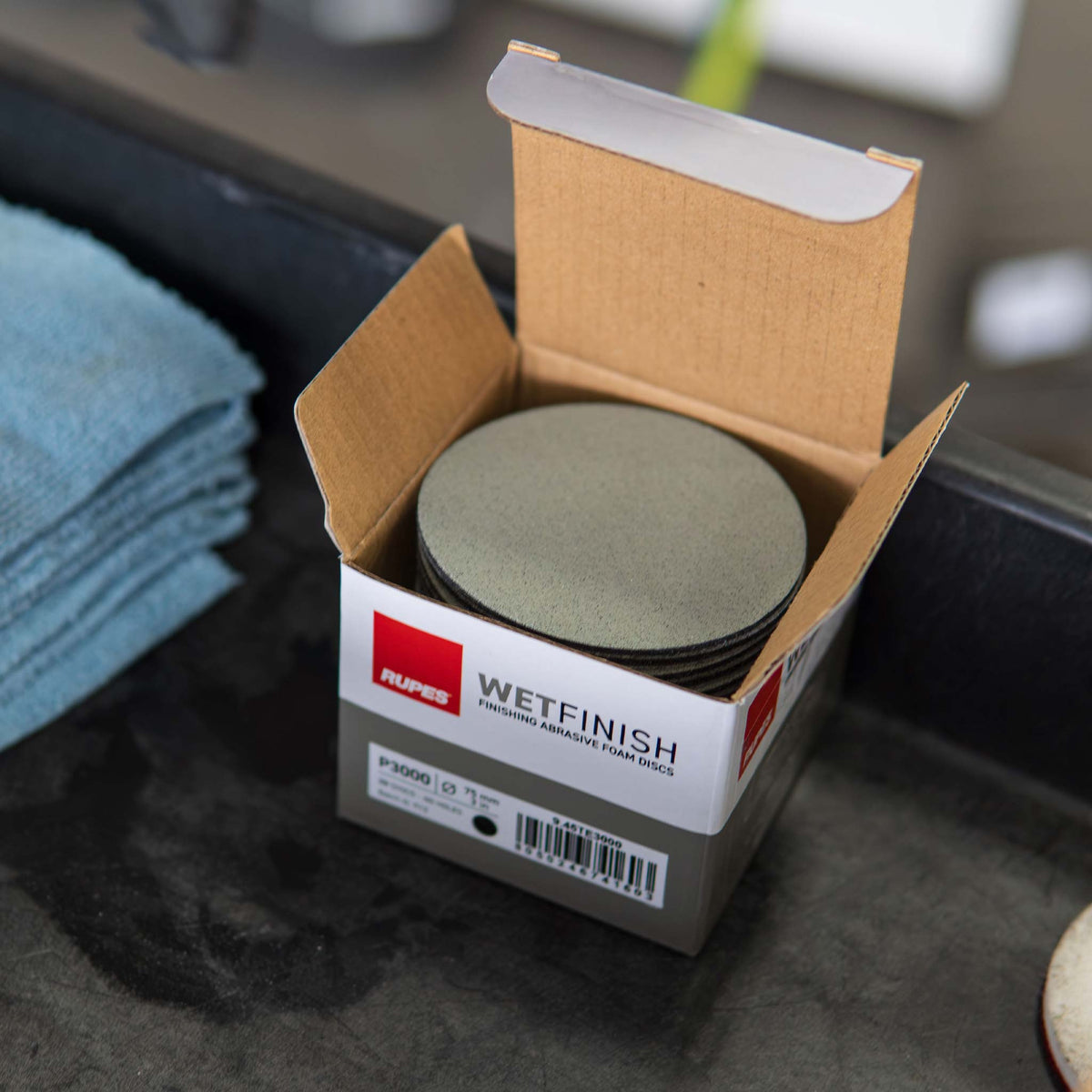 Open box of RUPES WetFinish P3000 discs beside microfiber towels on a detail cart