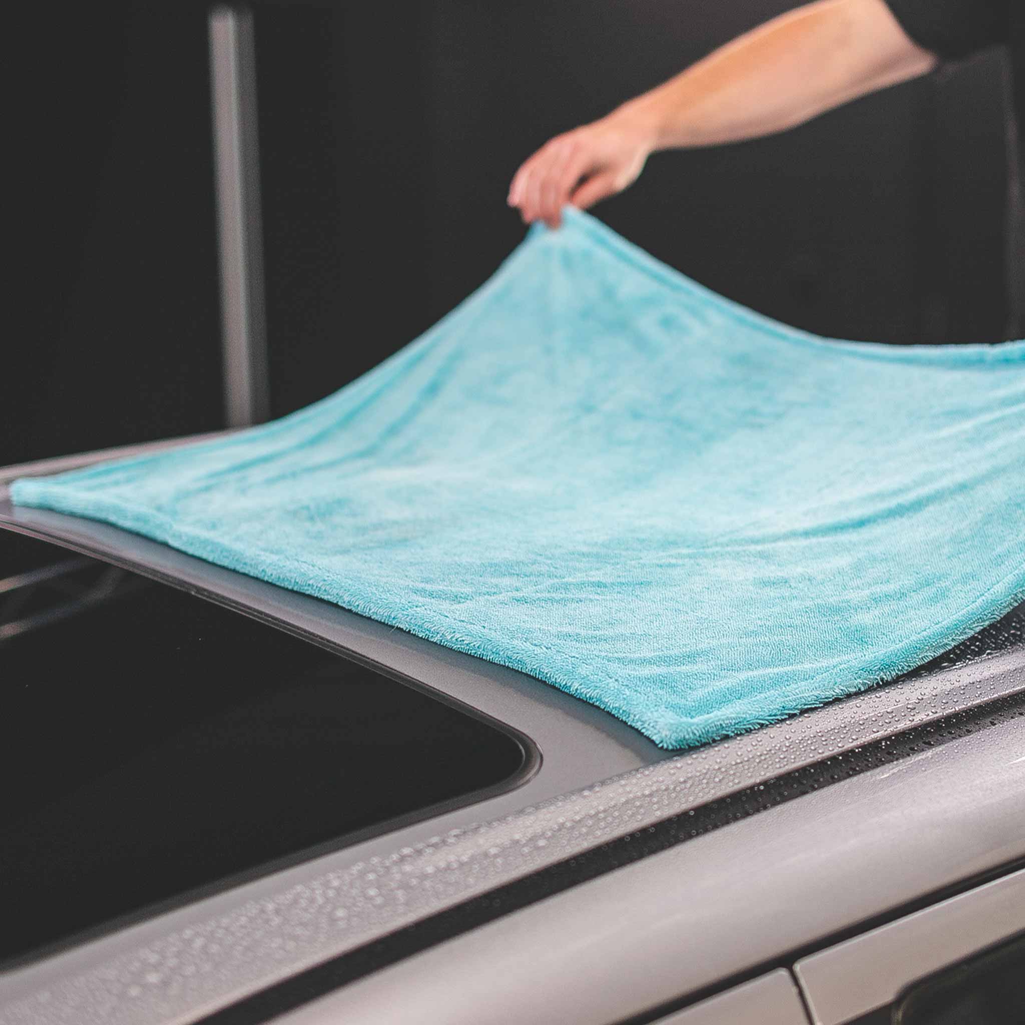 A person's hand is shown lifting a light blue microfiber towel over the roof of a silver car, with water droplets visible on the wet surface.