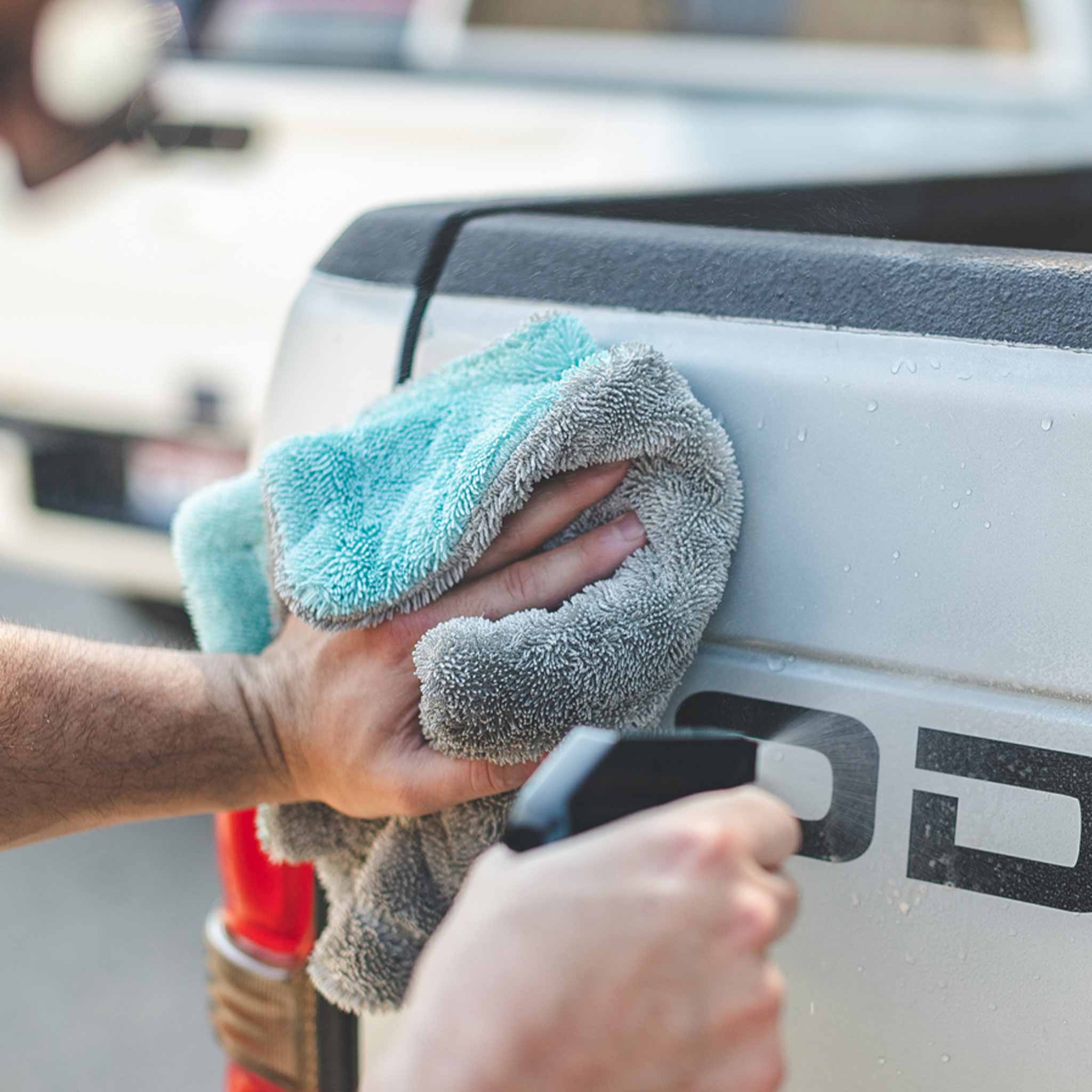 A close-up lifestyle shot of a person using a gray microfiber cloth to wipe down the side of a white pickup truck bed, with a spray bottle in hand.