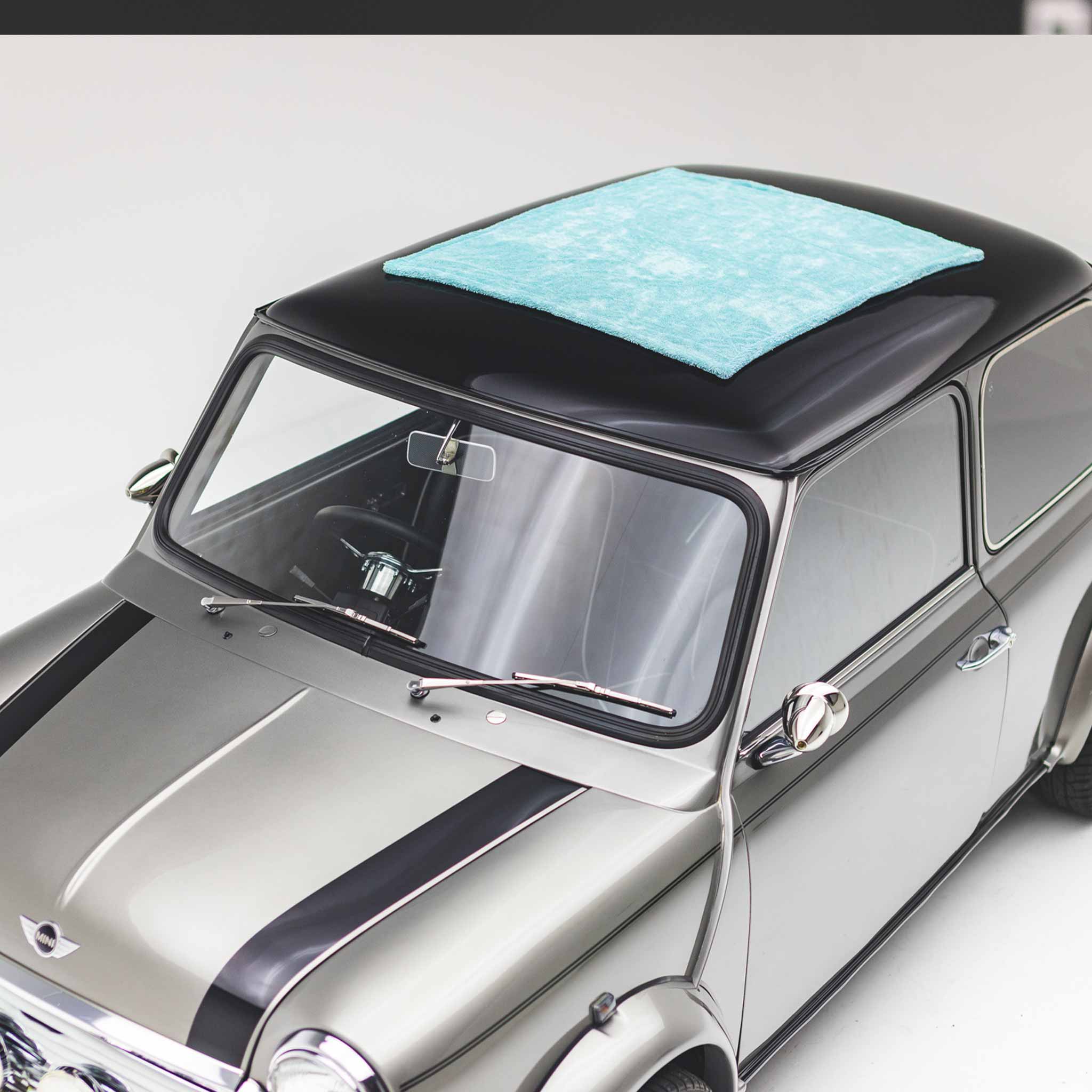 A silver classic Mini Cooper car with a black roof and a light blue microfiber towel placed on top, photographed in a clean studio setting.