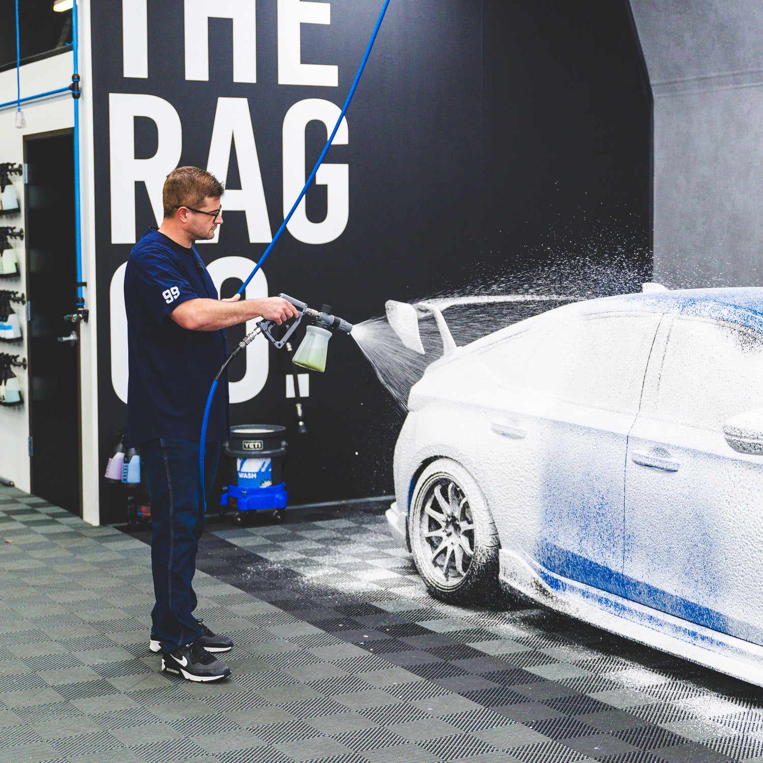 A man in a dark navy blue shirt and jeans washes a white car covered in foam inside a garage or detailing shop. A large black wall behind him has bold white text reading 'THE RAG'.