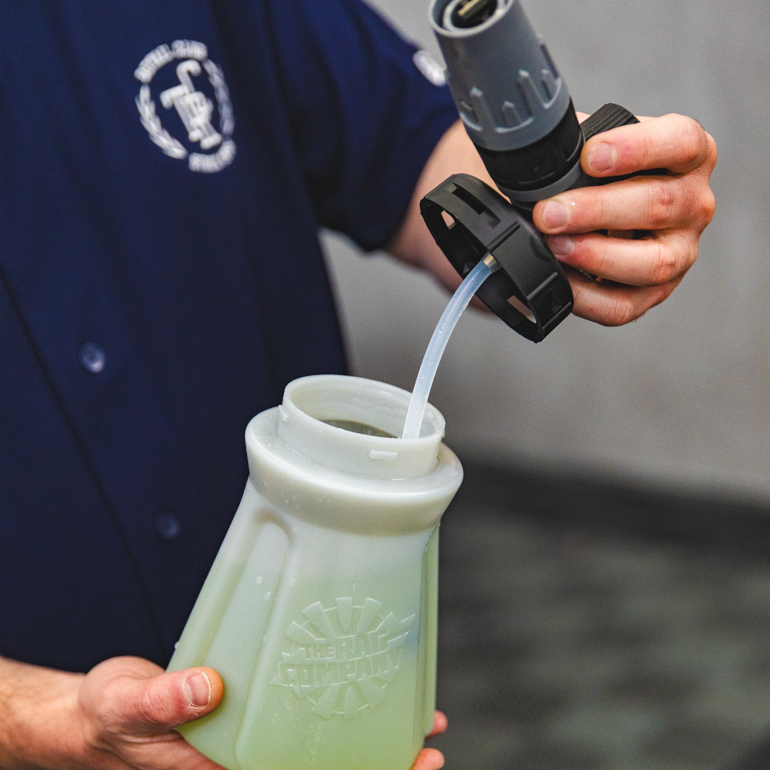 A person in a navy blue uniform is pouring water from a hose into a translucent grey plastic container filled with green soap. The person's hands and torso are visible, and the background is a blurred gray outdoor setting.