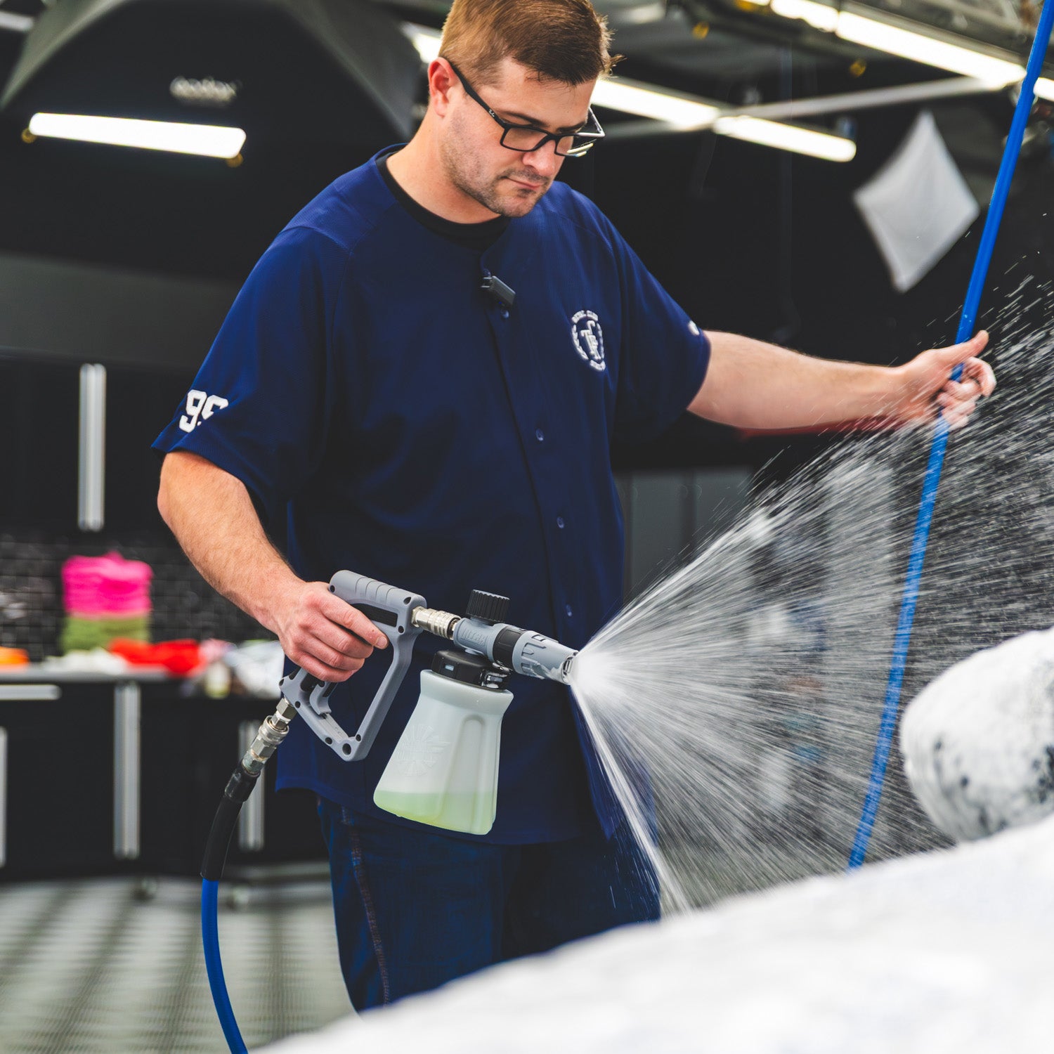 A man wearing a navy blue uniform sprays foam onto a white vehicle using a pressure washer wand in a garage setting.