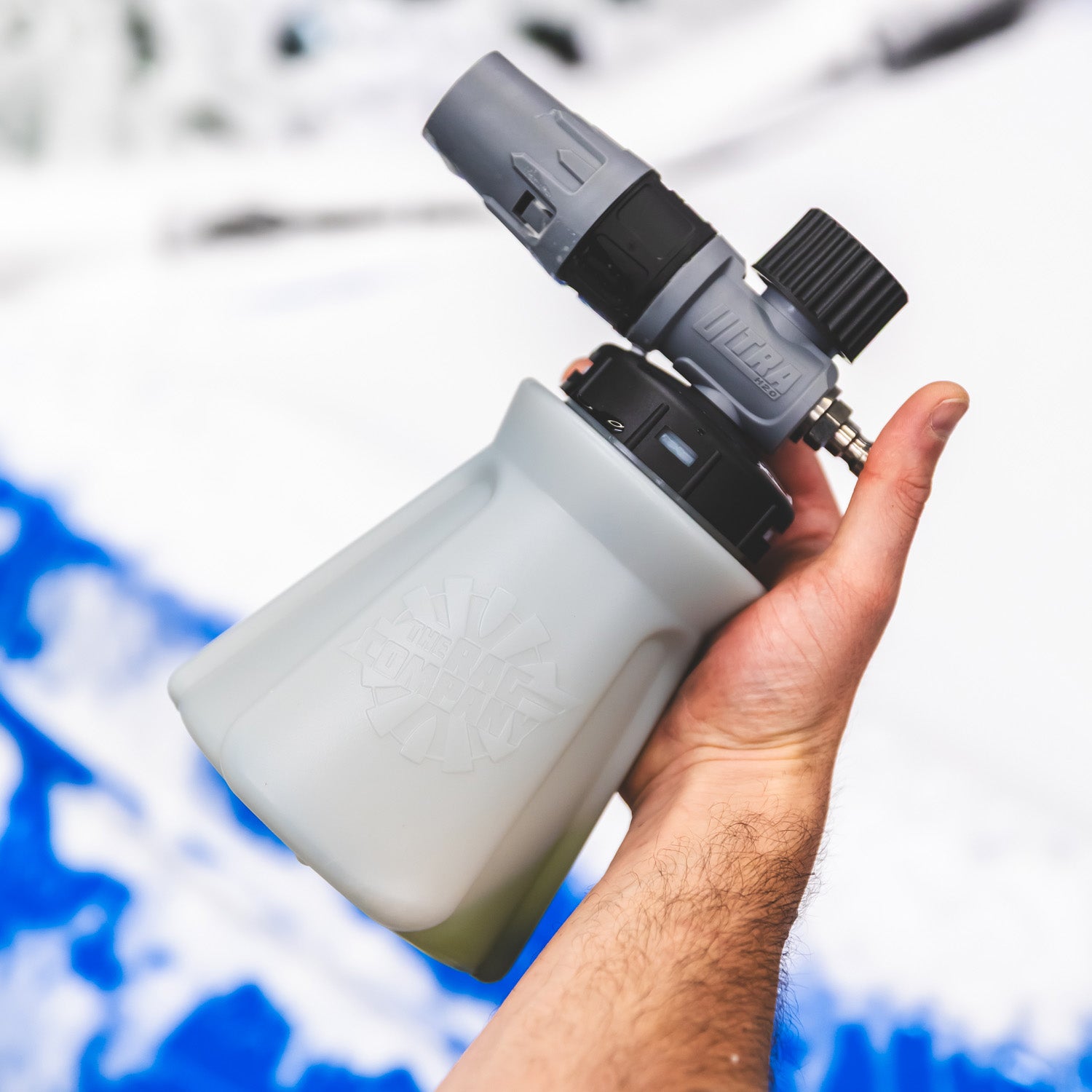 A hand holding a white and gray foam cannon attachment for a pressure washer, with a snowy outdoor background.