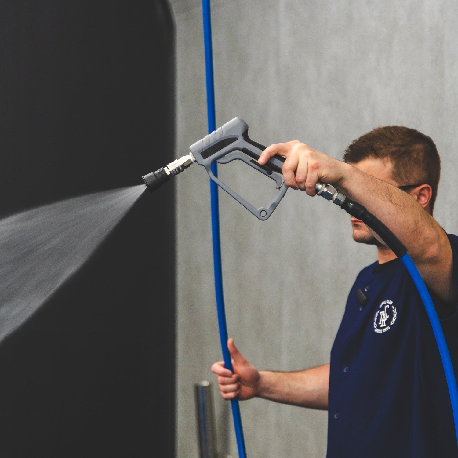 A man in a navy blue uniform sprays water from a high-pressure hose wand against a dark wall in a clean service environment.
