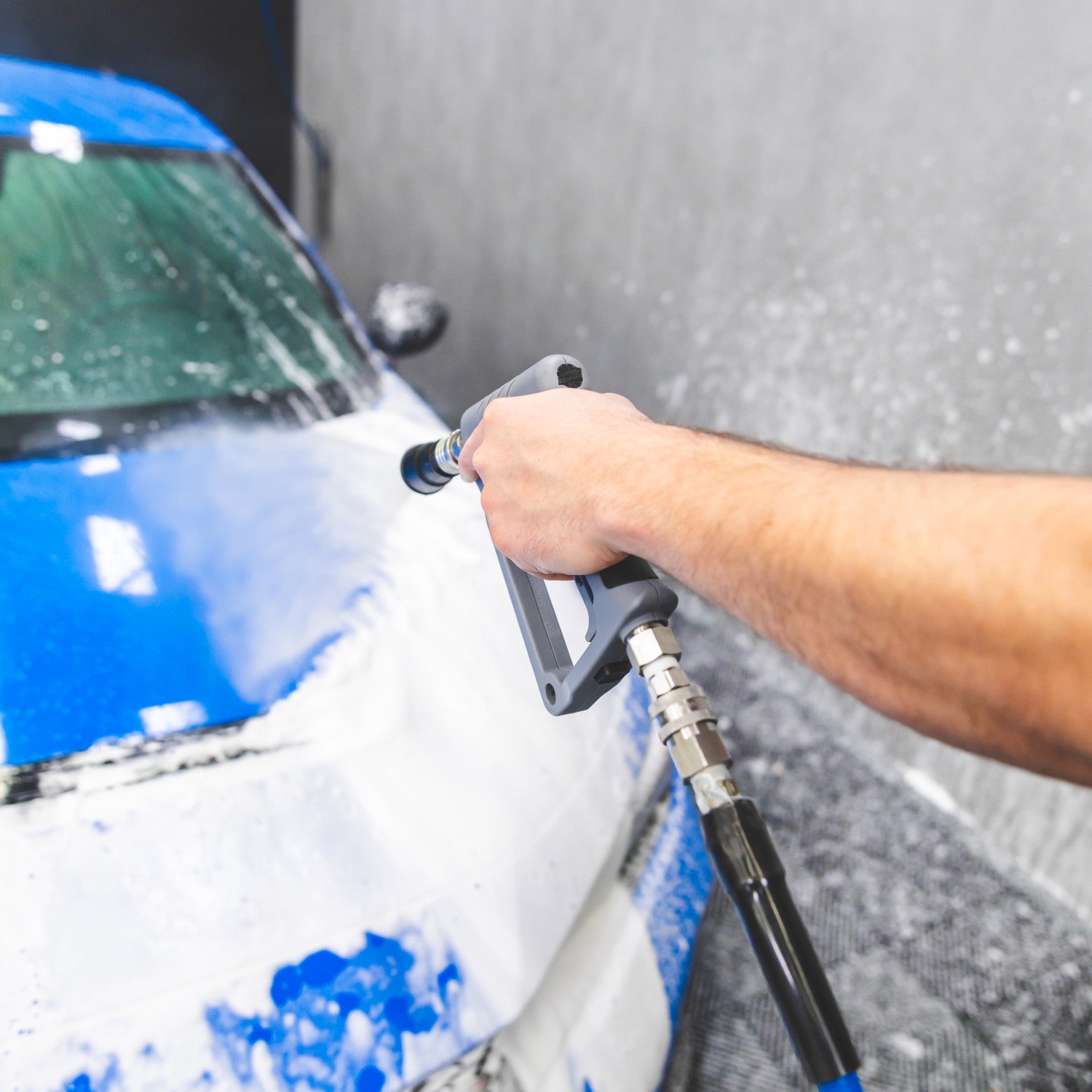 A close-up action shot of a person's arm holding a high-pressure water hose and spraying soap lather onto the front hood of a white car with a blue front bumper.