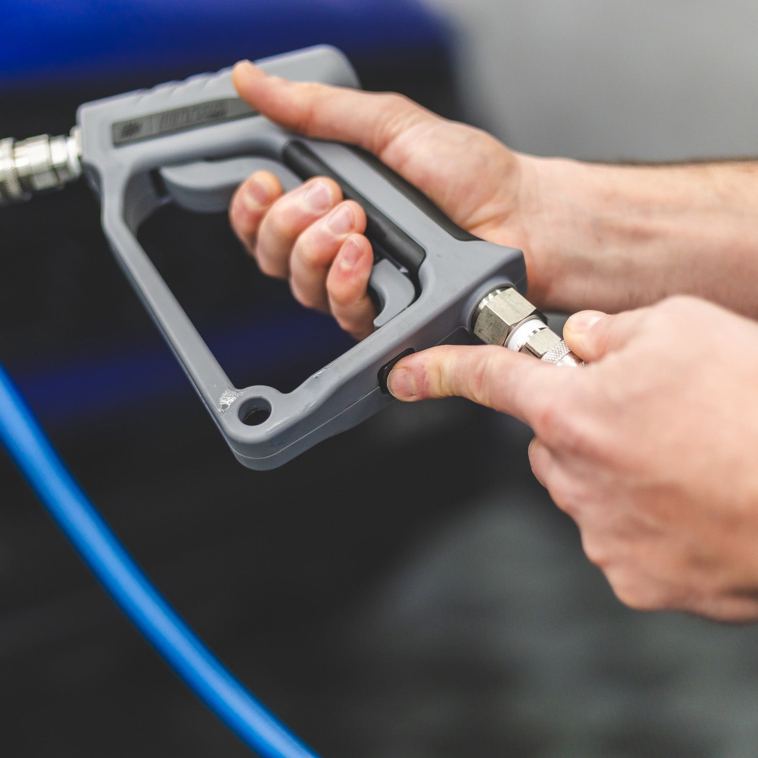Close-up of a person's hands holding and pointing to a gray high-pressure water gun attachment with a metal connector, connected to a blue hose against a dark blurred background.