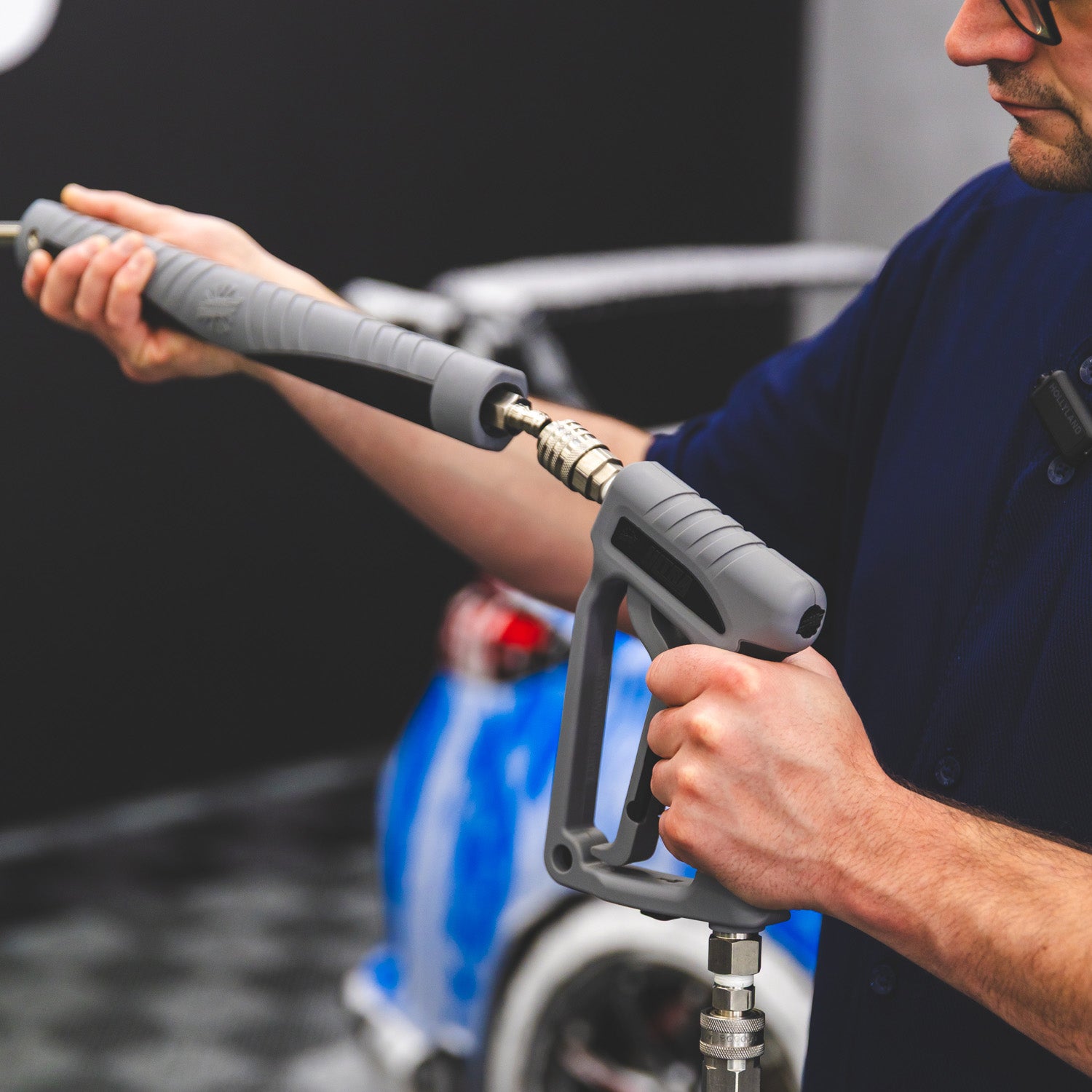A close-up of a person holding a gray high-pressure water spray gun attachment, with a blue and white vehicle blurred in the background.