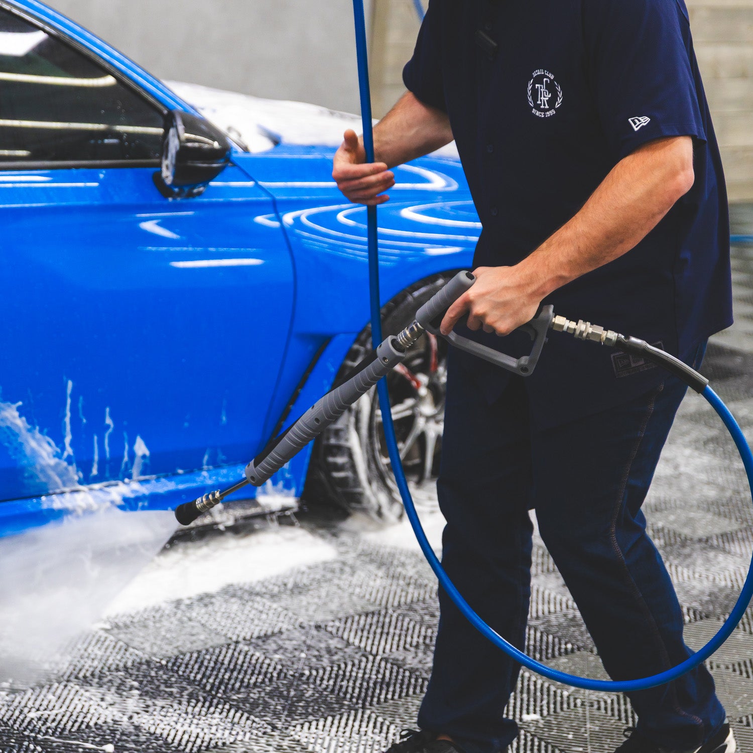 A person in a navy blue uniform uses a high-pressure water hose to wash foam off a bright blue car at an outdoor car wash.