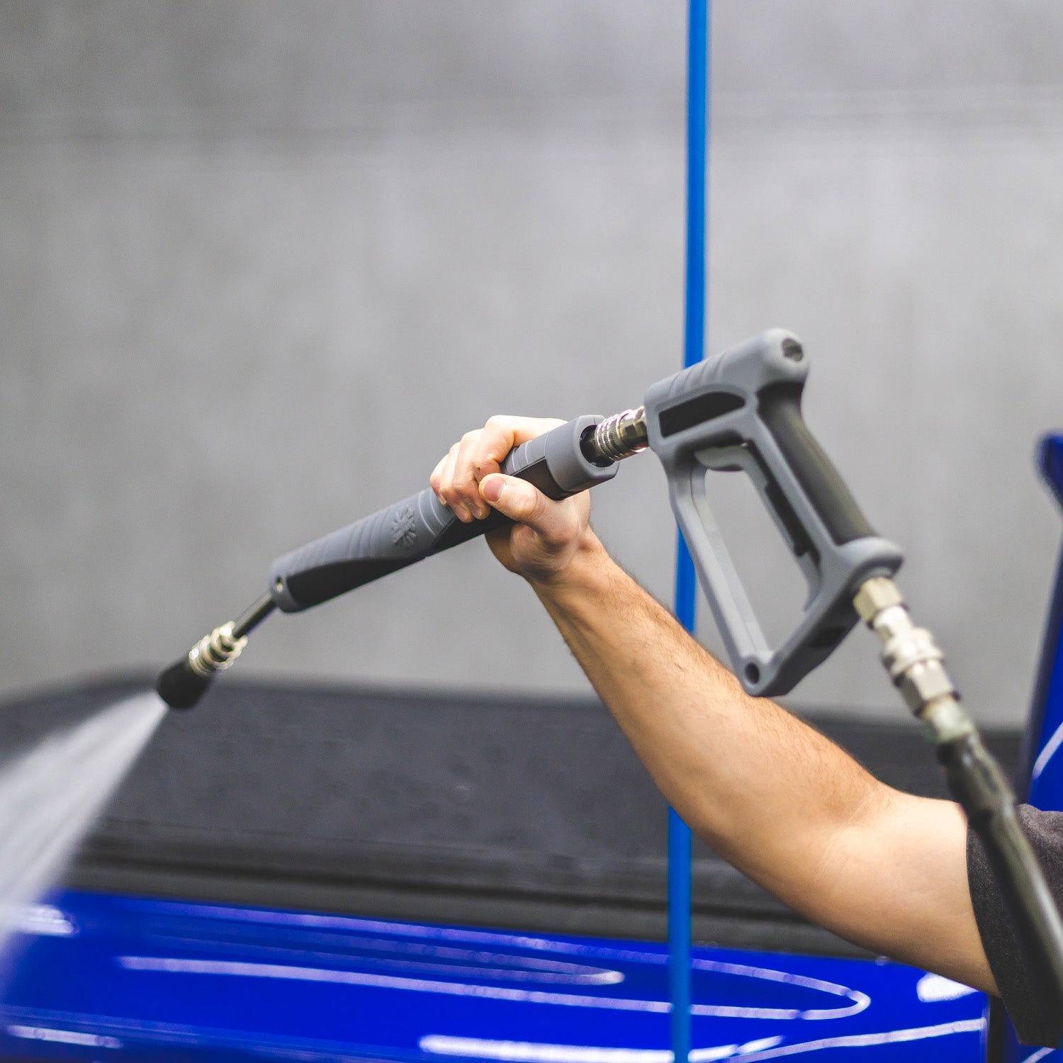 A close-up of a person's arm holding a gray high-pressure water spray wand, spraying water onto a glossy blue vehicle in a gray indoor setting.