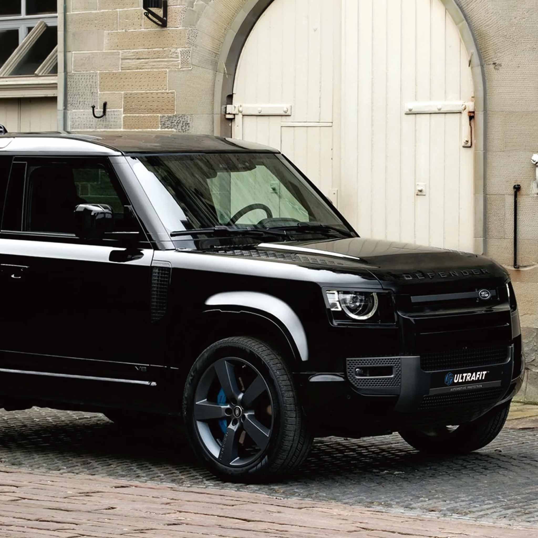 A black PPF custom Land Rover Defender SUV parked on a cobblestone street in front of a stone building with a large white garage door. 
