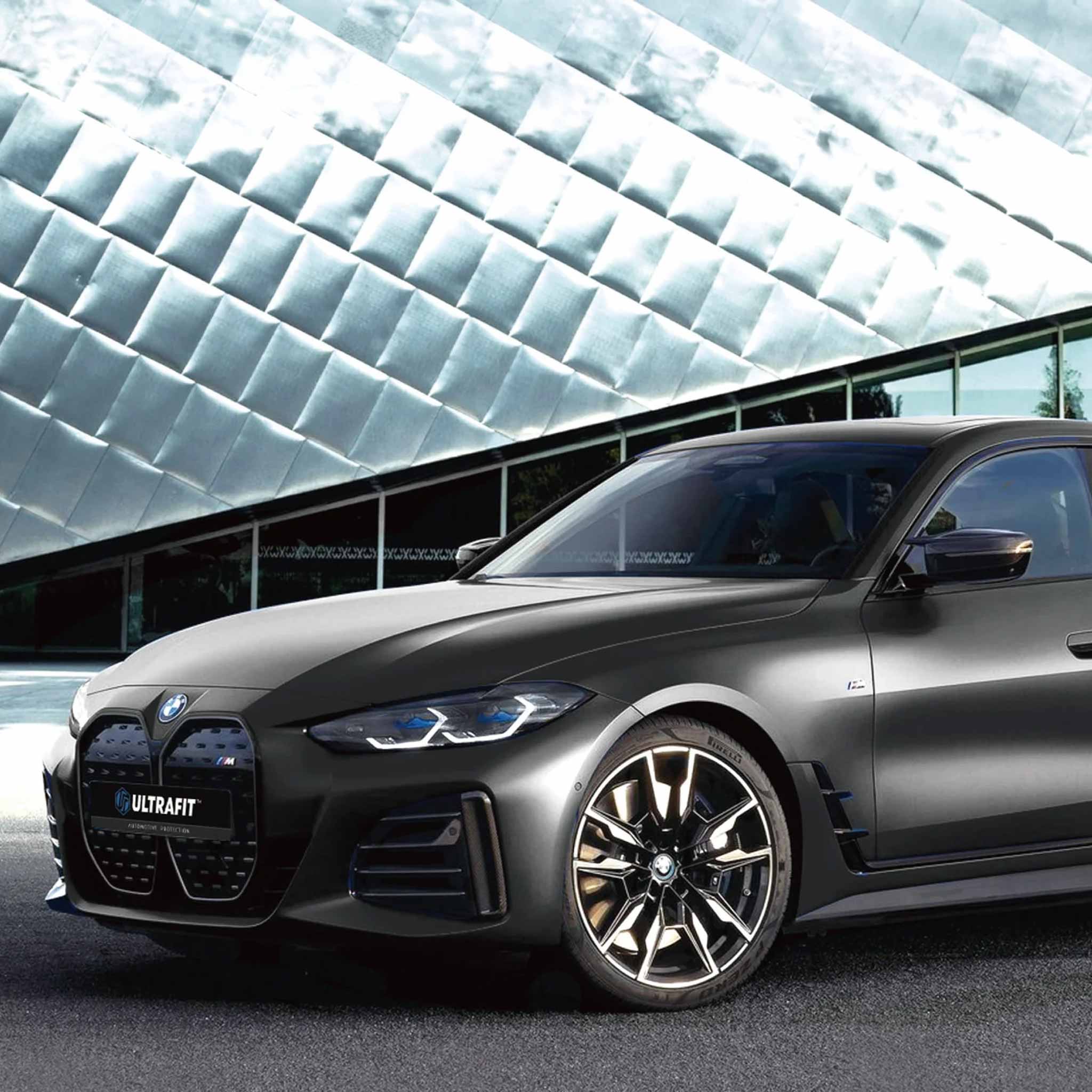 A black matte BMW M4 Competition with a UCRafT front grille badge, parked in front of a modern glass building with a distinctive quilted metallic facade.
