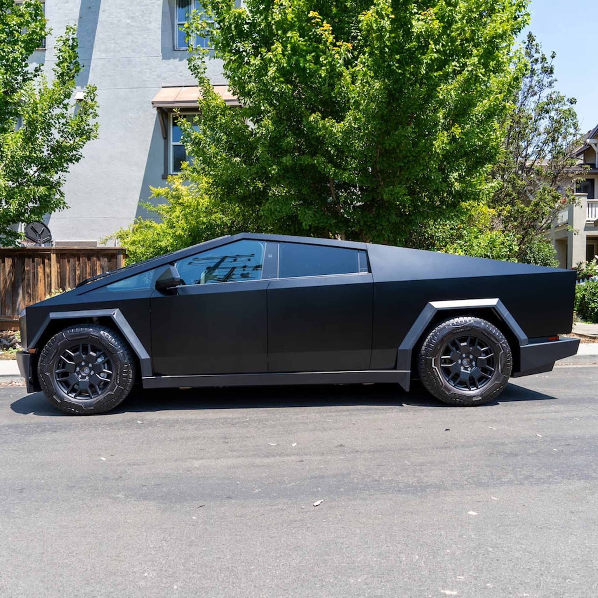 A matte black Tesla Cybertruck parked on an asphalt street in front of a house with green trees.