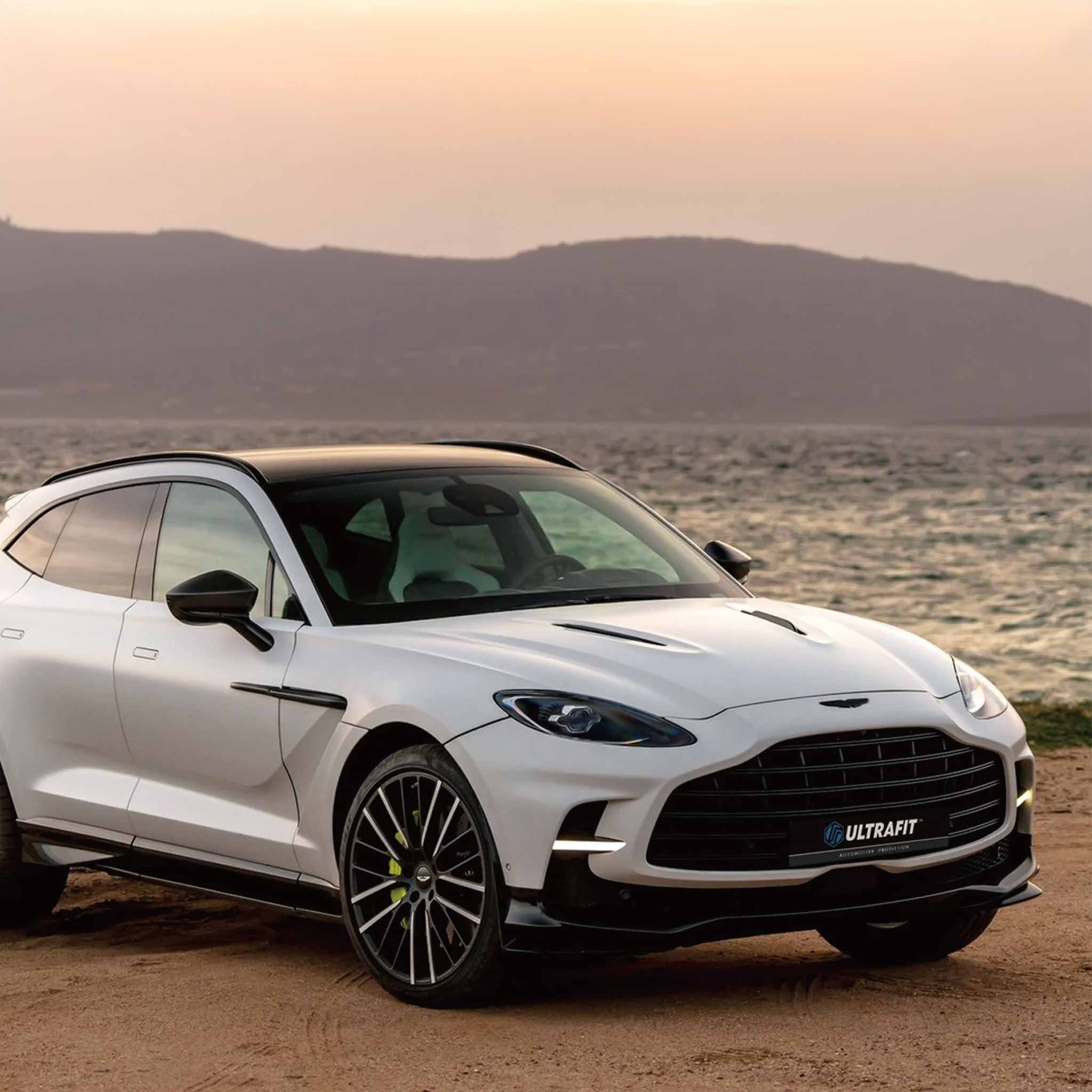 A white Aston Martin DBS Ultra is parked outdoors on a sandy shore with water and hazy hills in the background. The car has matte ppf on it features a black grille, black side mirrors, black wheels, and a small blue 'ULTRAFIT' badge on the front grille.