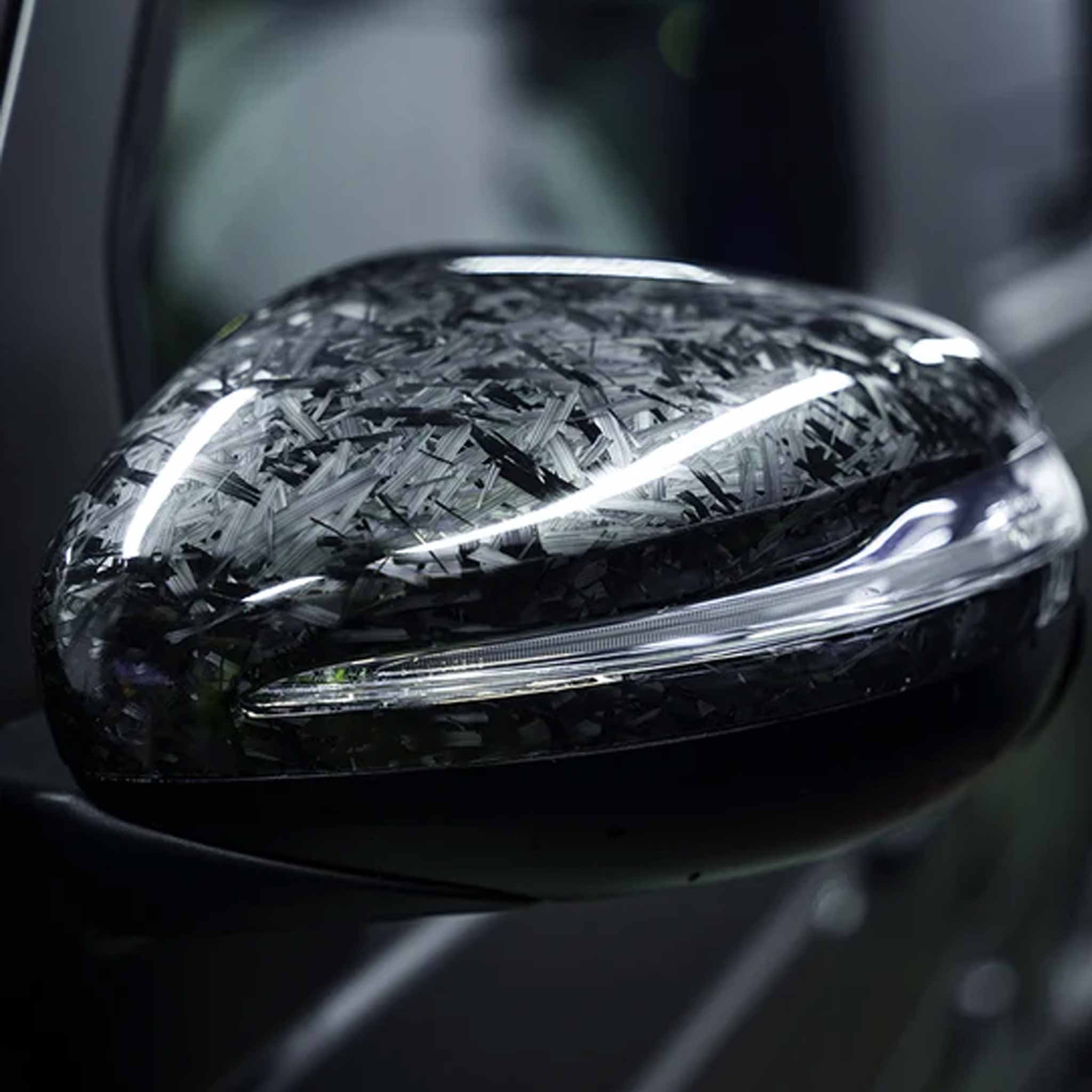 A close-up of a glossy black car side mirror housing covered in a textured, metallic carbon-fiber-like protective wrap, with a clear LED light strip visible along the front.