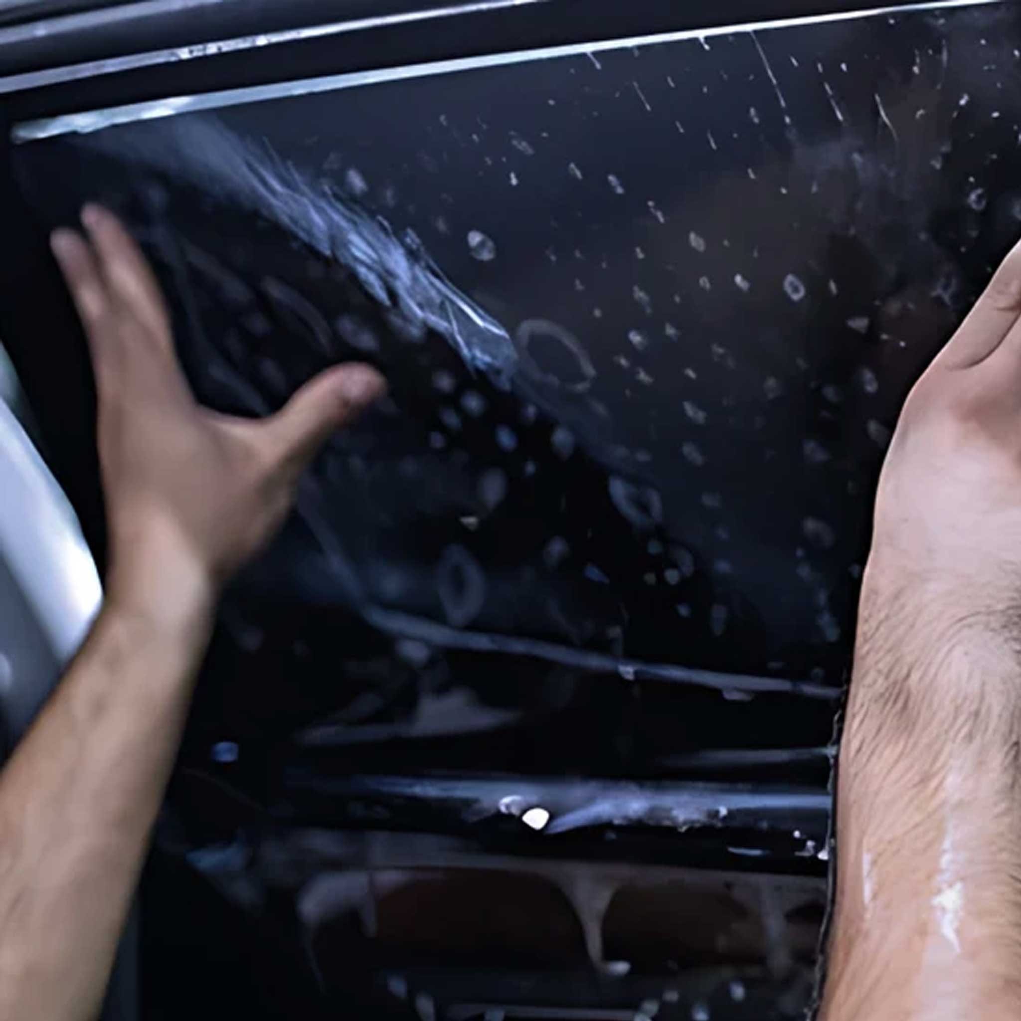 Close-up of a person's hands applying a glossy, water-droplet-covered film or liner to a dark car window.