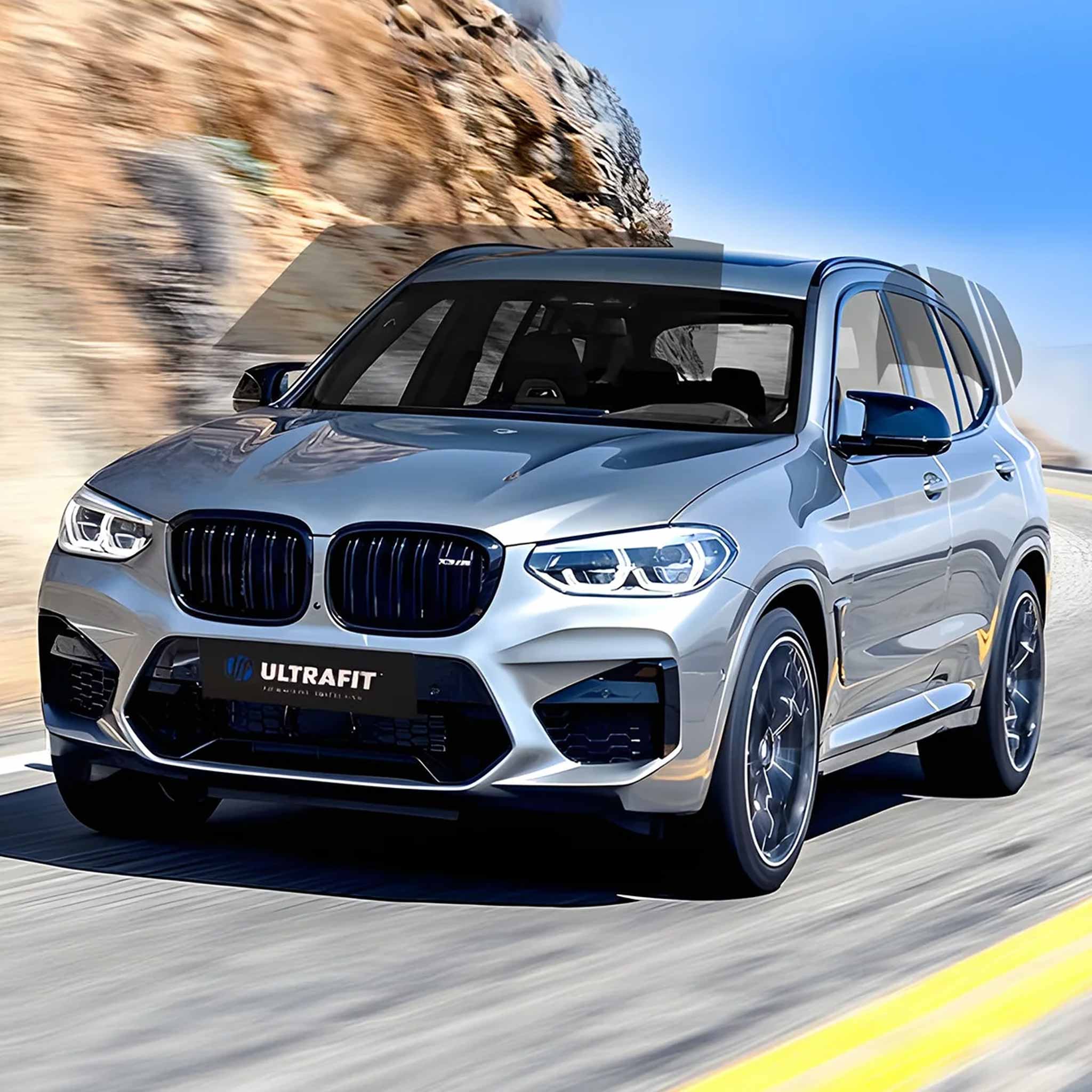 A silver BMW X3 M SUV with an ULTRAFIT M carbon fiber body kit, shown in motion on a winding road with a blurred rocky hillside background.