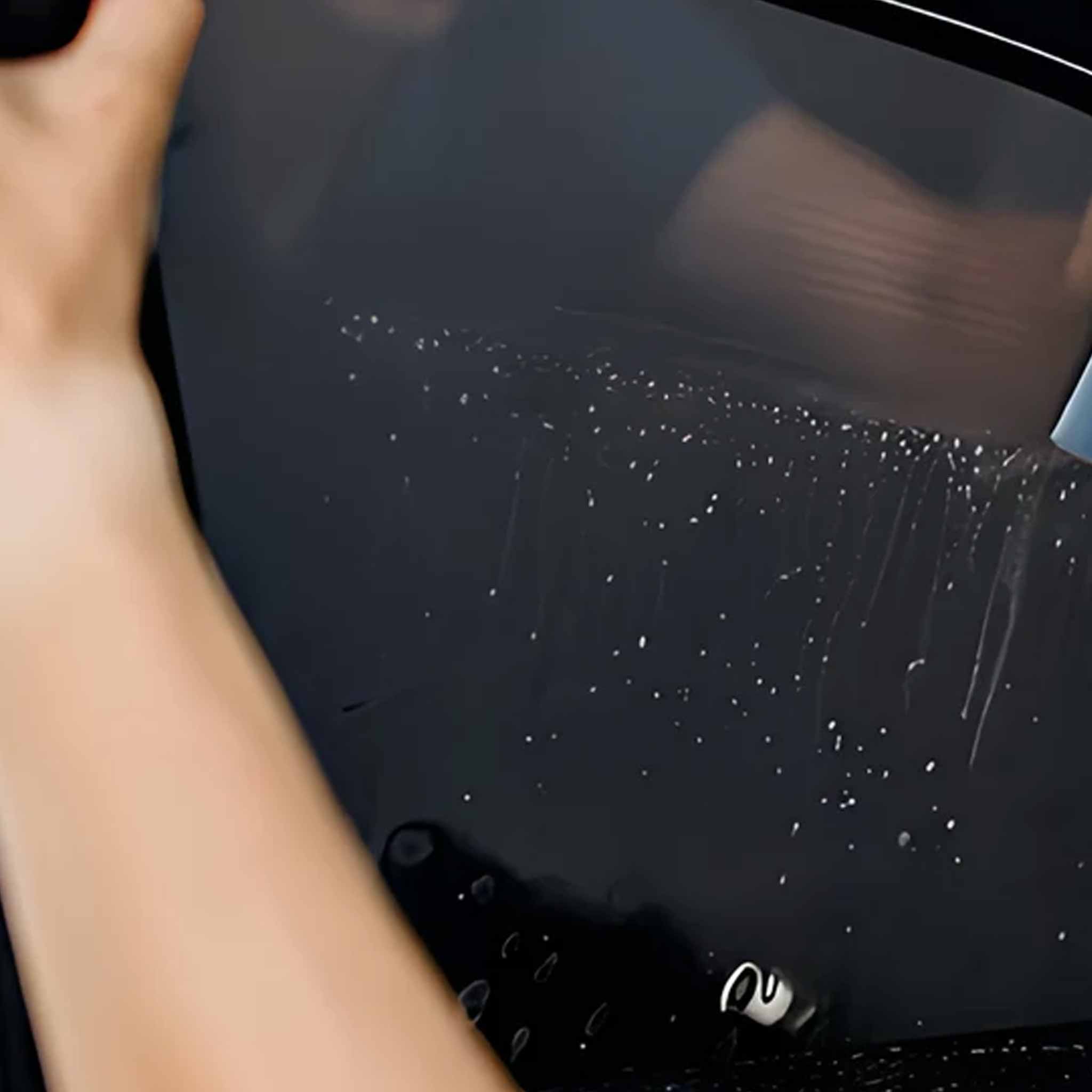 A close-up of a hand applying or smoothing a clear, water-droplet-window film onto a dark tinted car window.
