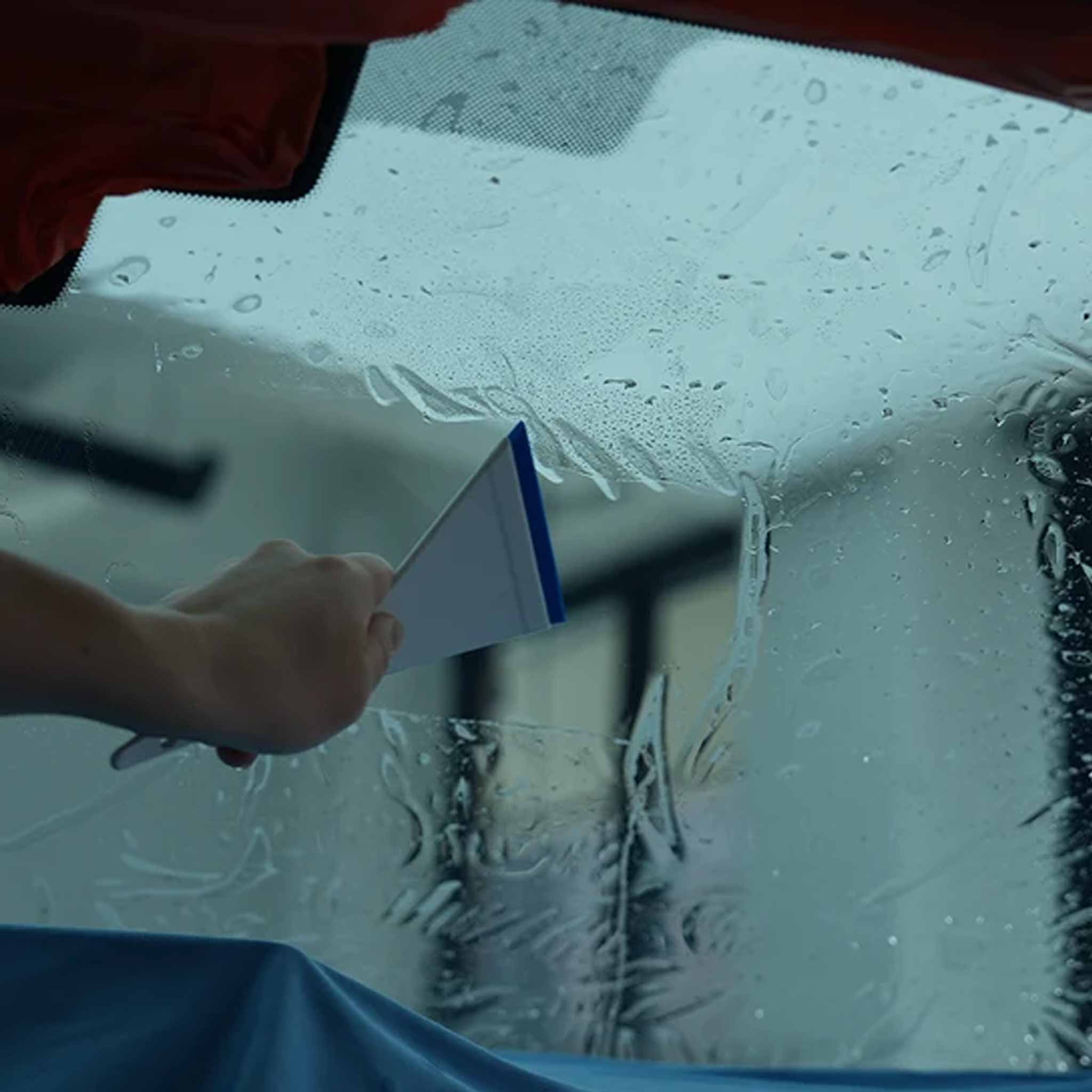 A hand using a triangular squeegee to clear water from a car windshield applying vivid signature window tint